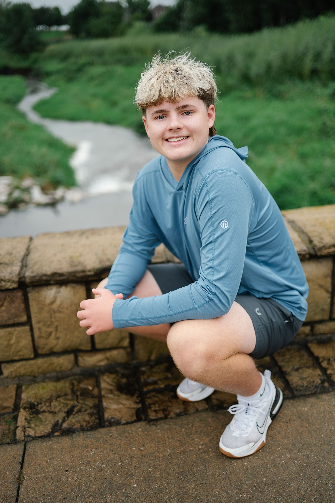 Smiling young man with blonde hair crouching on stone wall wearing blue hoodie and shorts near a stream.