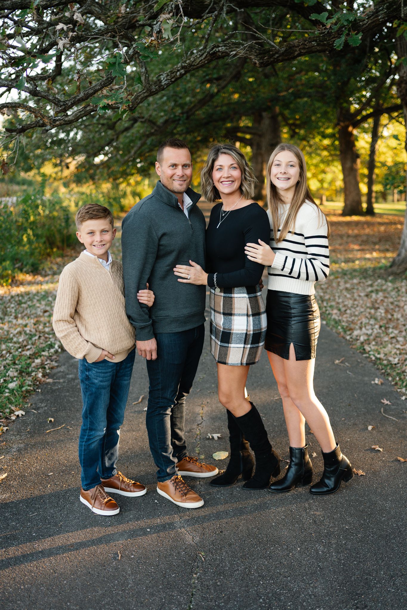 Family of four posing together on a tree-lined park path in autumn, with a boy, a man, a woman and a teenage girl smiling.