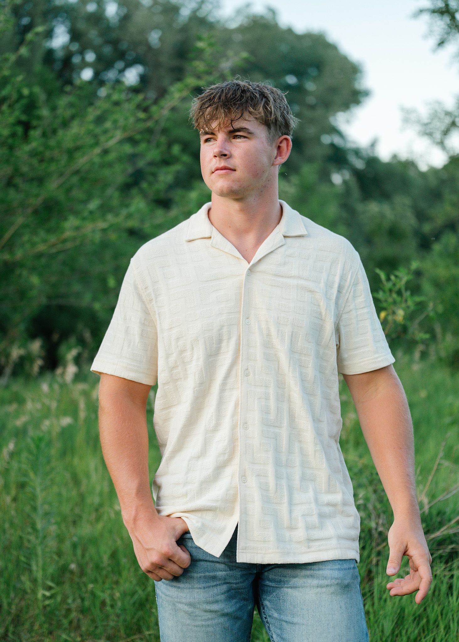 Young man in cream shirt and jeans standing in lush green field during golden hour outdoor photo session for Ankeny family photographer.