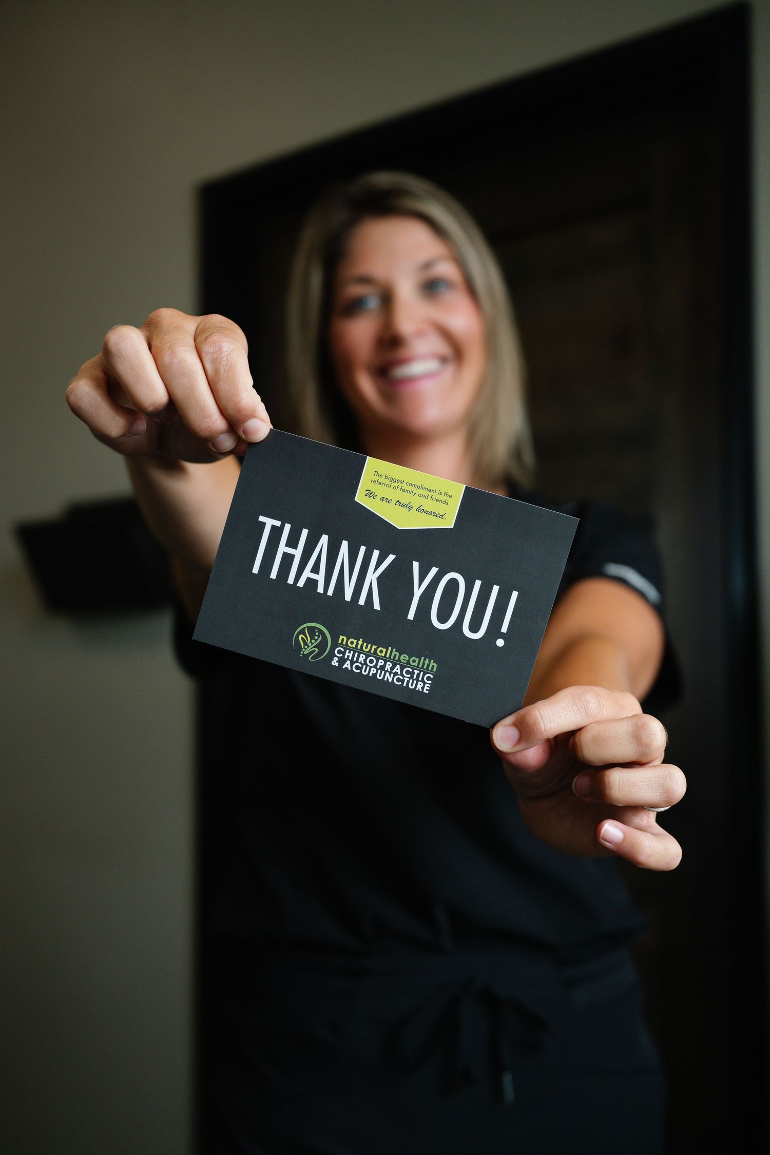 Smiling woman in scrubs holding a black Thank You card from Nature Health Chiropractic and Acupuncture clinic.