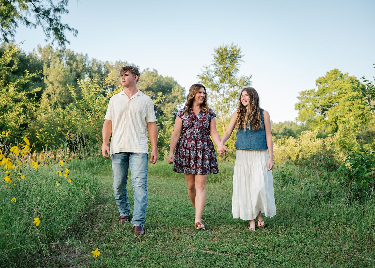 Three people walk hand-in-hand through a lush green meadow with wildflowers on a sunny day posing for Ankeny family photographer.