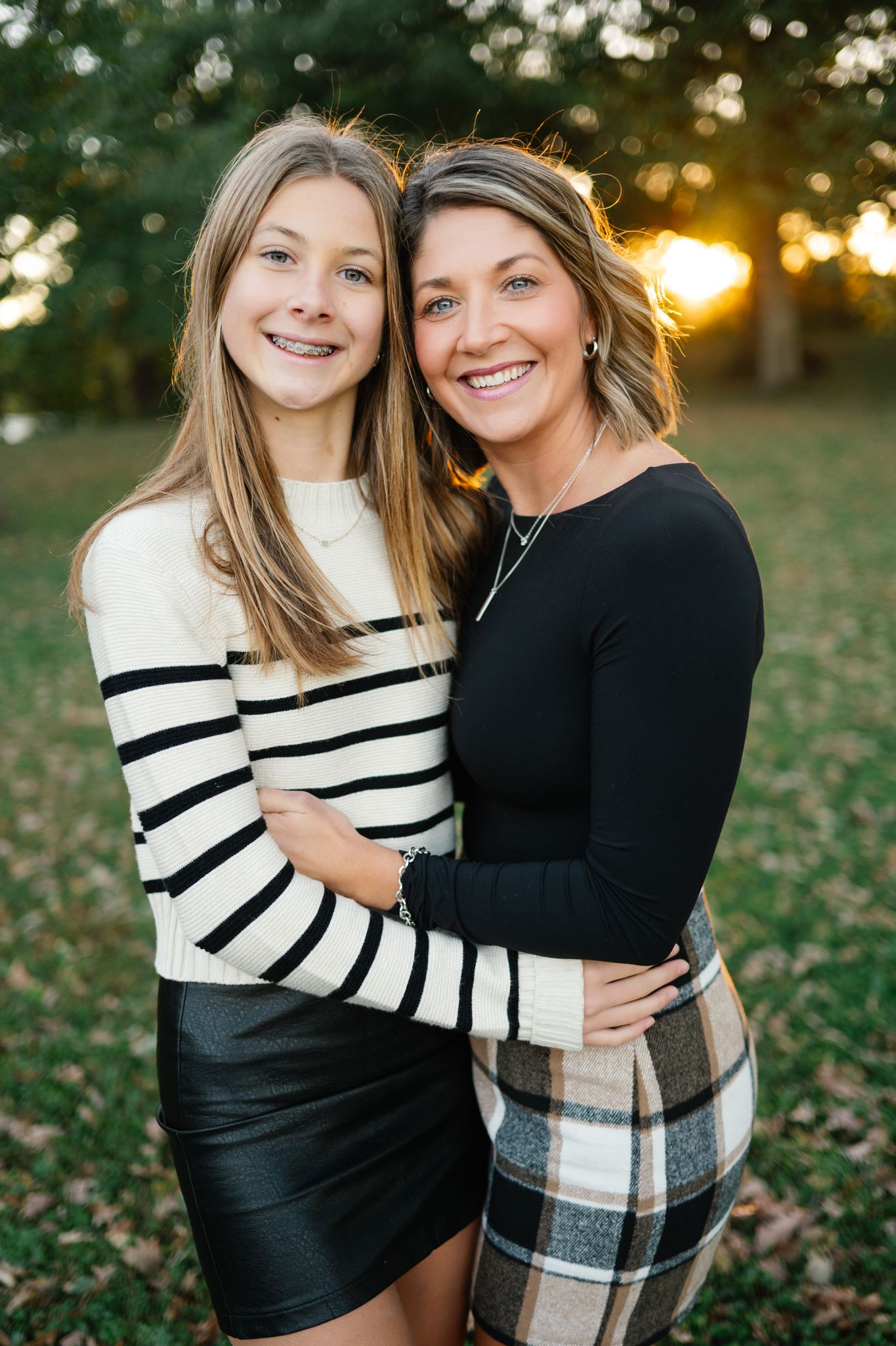 Smiling girl in a striped sweater and leather skirt hugs a woman in a black top and plaid skirt outdoors at golden sunset.