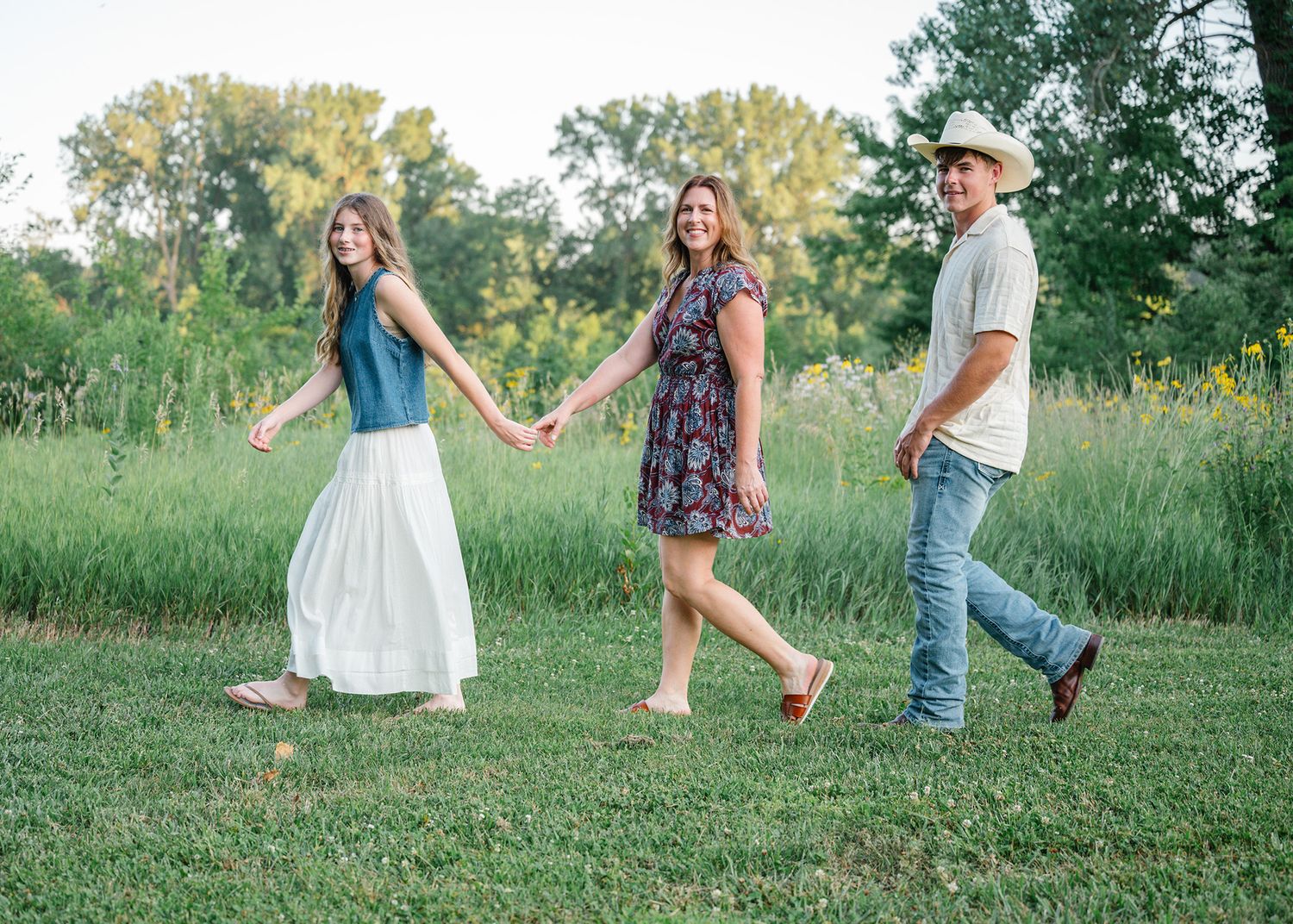 Three people holding hands and walking through a lush green field surrounded by trees on a sunny day posing for Ankeny family photographer.
