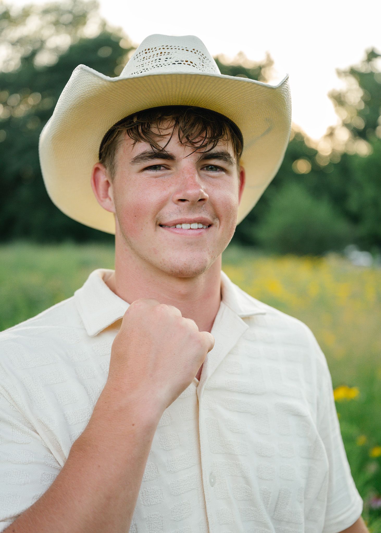 Young man in a straw cowboy hat smiling in a sunny field of wildflowers, wearing a cream polo shirt posing for Ankeny family photographer.