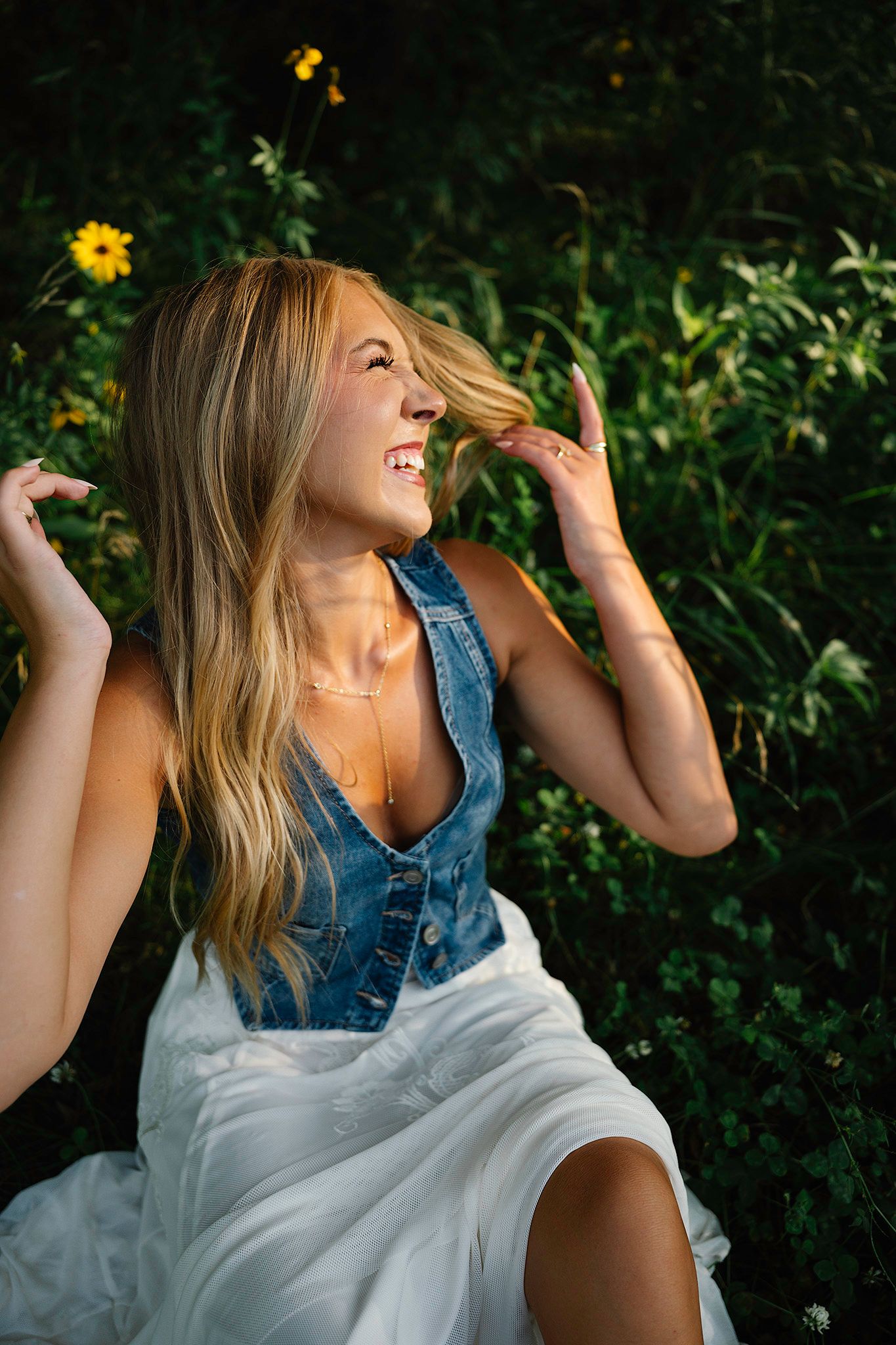 Smiling blonde woman in denim vest and white skirt sitting among wildflowers in golden sunlight.