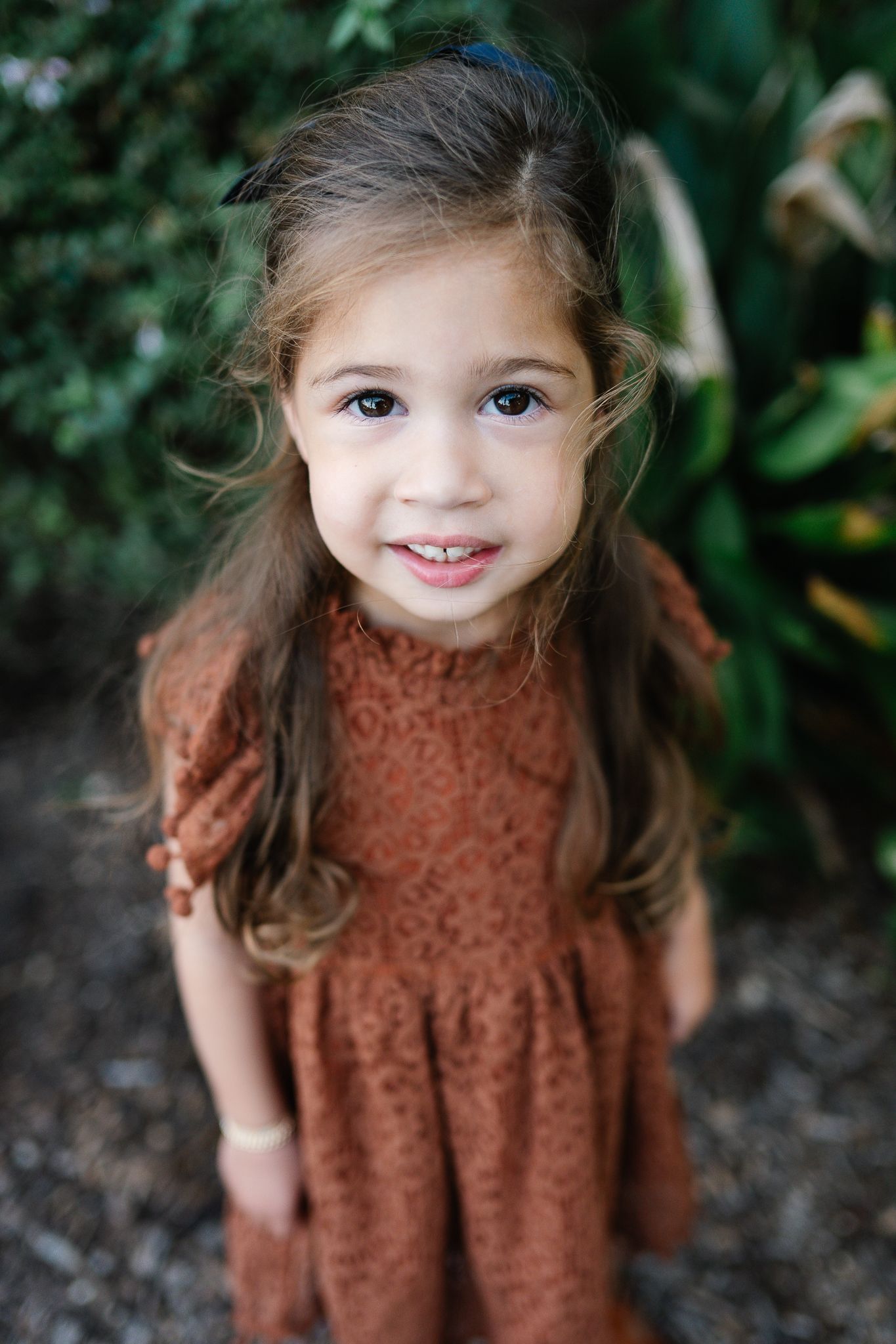 Smiling young girl with long brown hair wearing a rust-colored lace dress, standing outdoors in front of green leafy plants.