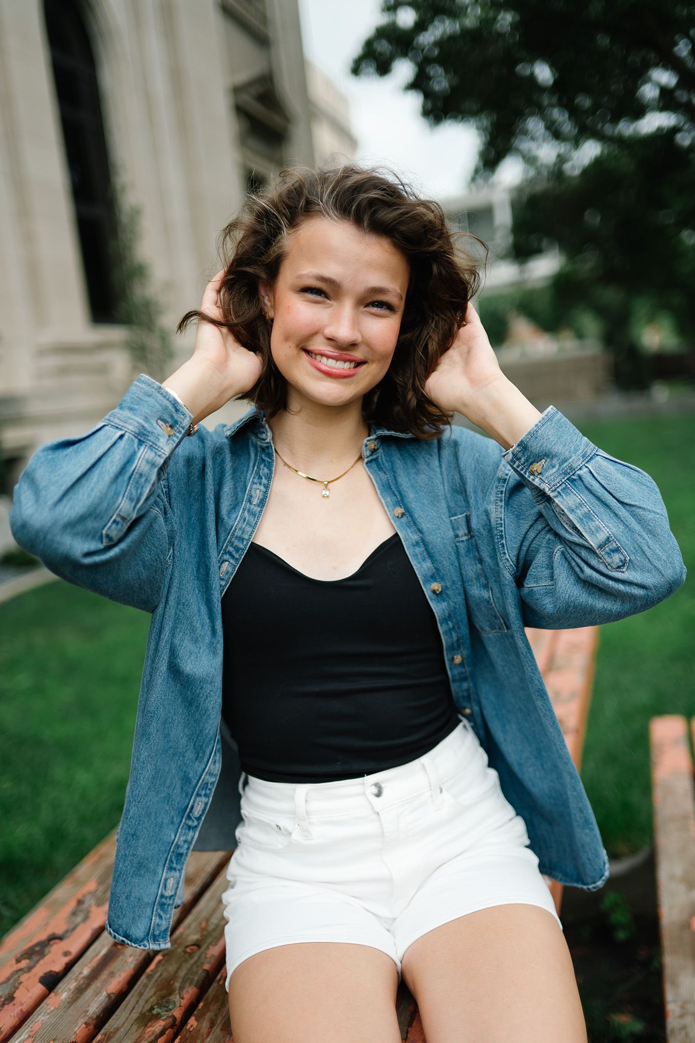 Young woman smiling outdoors, wearing denim jacket, black top, and white shorts, sitting on a bench with hands in curly hair.