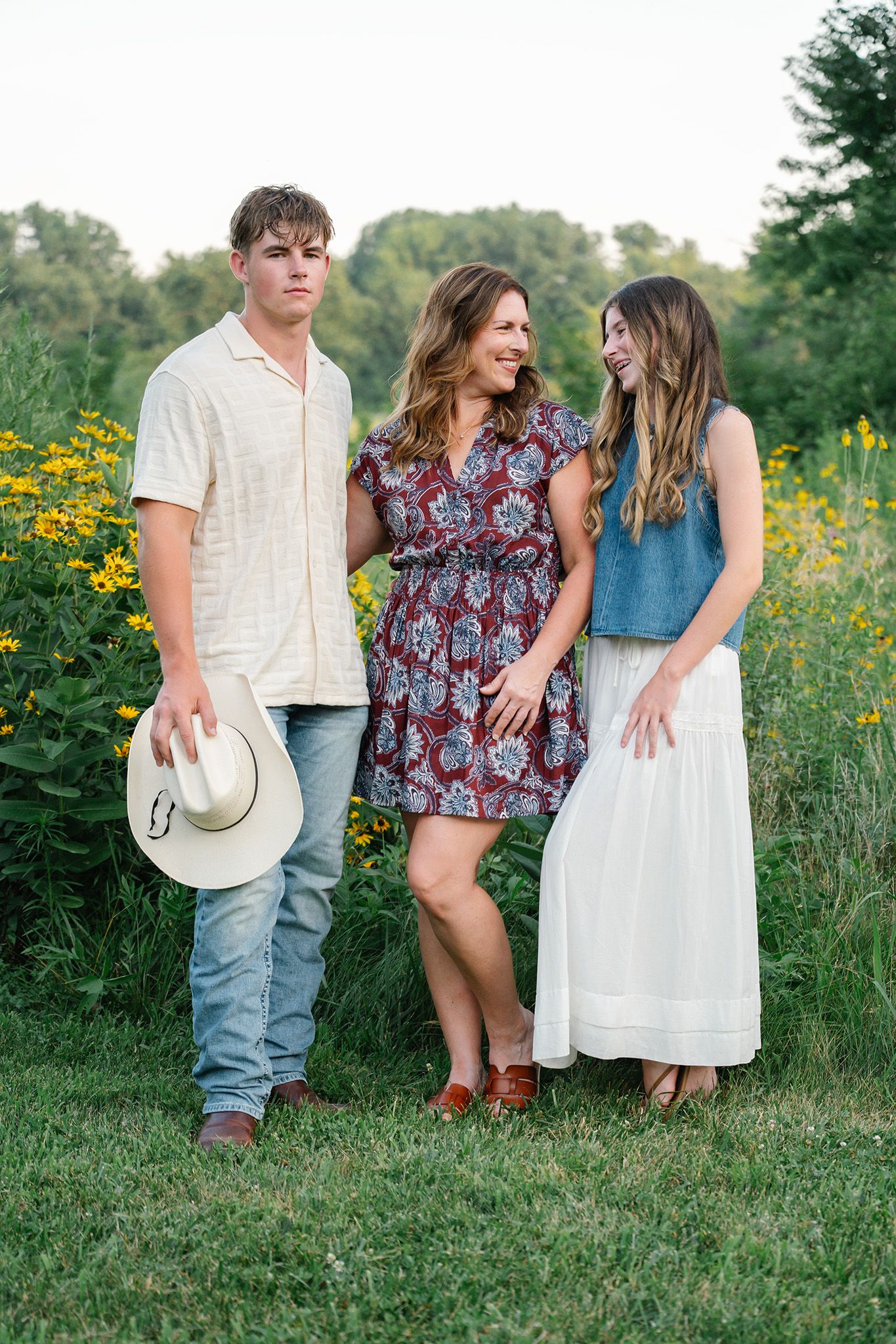 A mother poses with her teenage son and daughter in a sunny field of yellow wildflowers for Ankeny senior photographer.