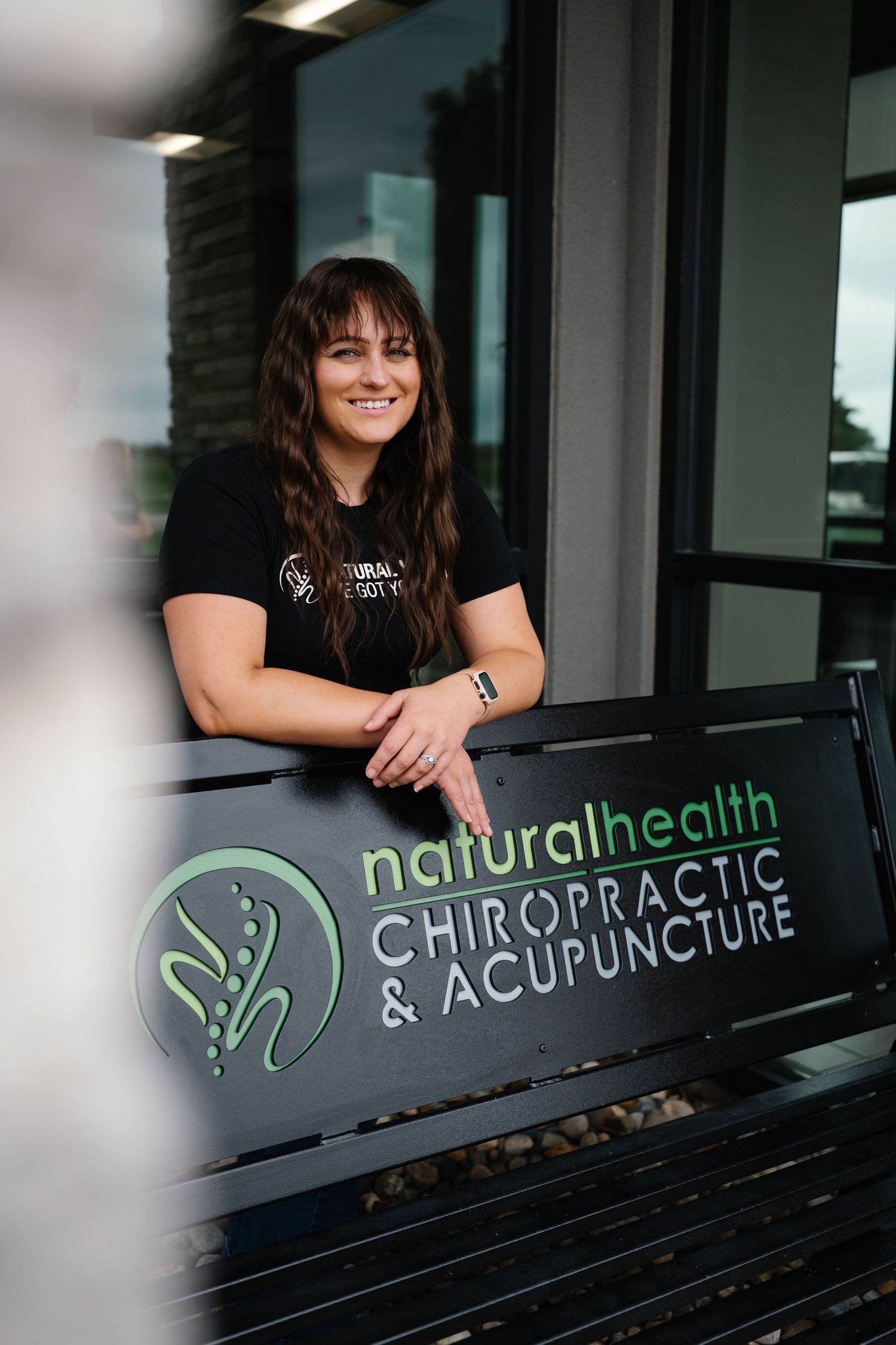 Woman smiling beside a Natural Health Chiropractic & Acupuncture sign outside a modern clinic.
