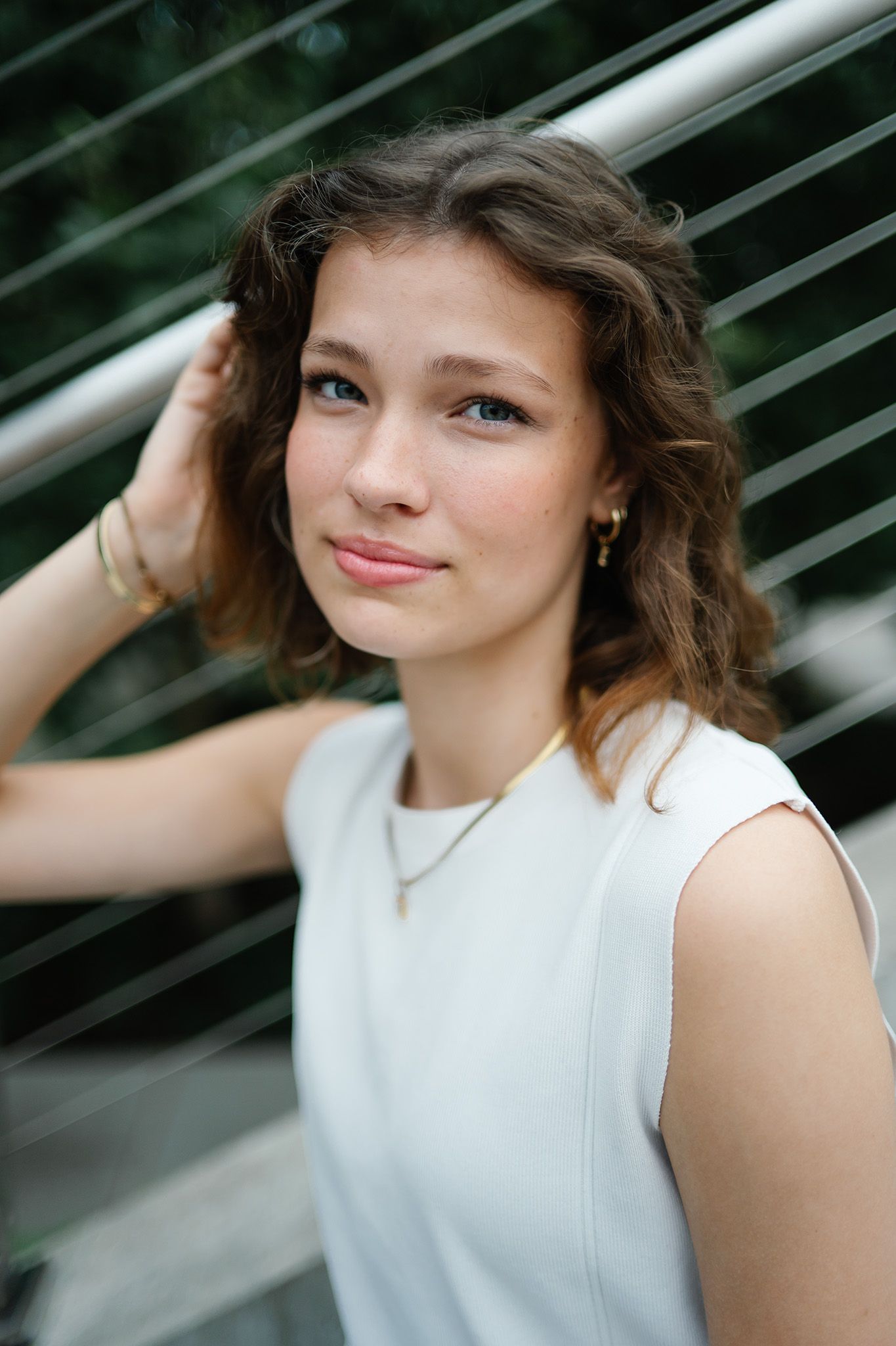 Young woman with curly brown hair wearing a white sleeveless dress and gold jewelry, posing near metal railings outdoors.