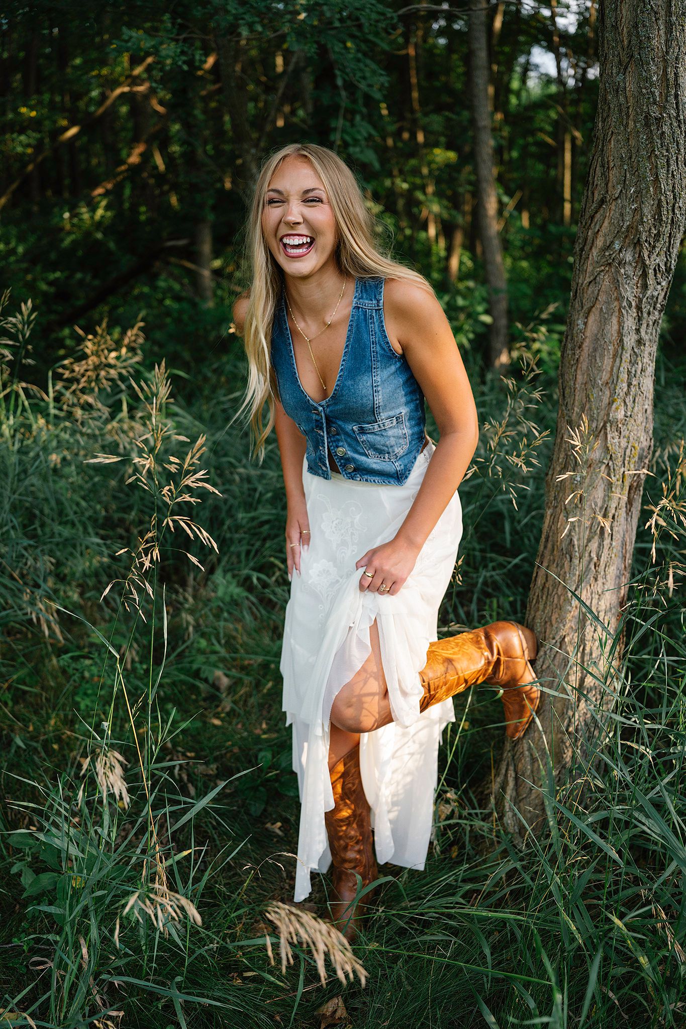 Laughing blonde woman in denim vest, white skirt, and boots posing by a tree posing for Des Moines senior photographer