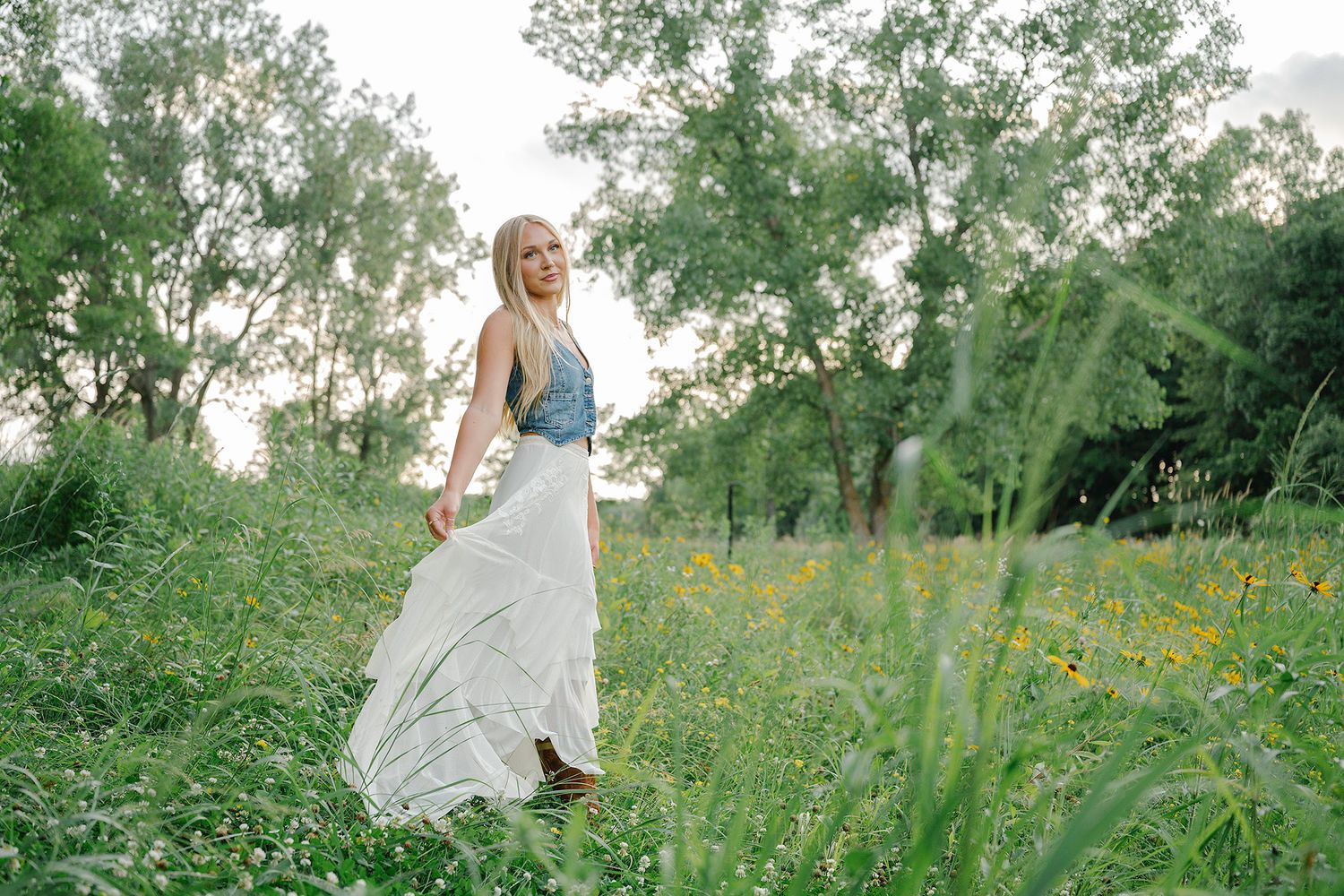 Blonde woman in white maxi skirt and denim vest walking through a lush green meadow with wildflowers.