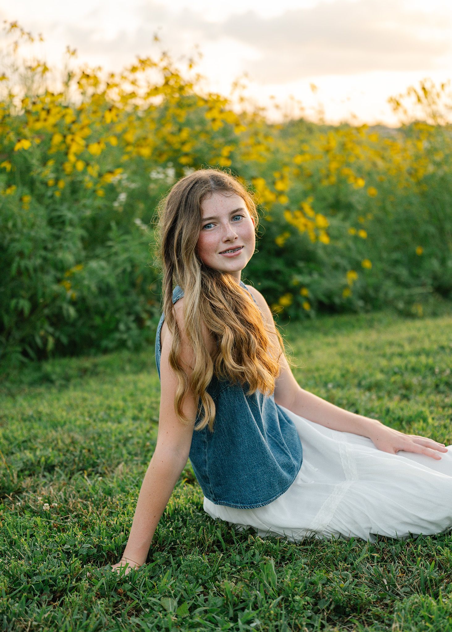 Young girl with long hair sits on green grass in a white skirt and denim top, with yellow wildflowers behind, posing for Ankeny photographer