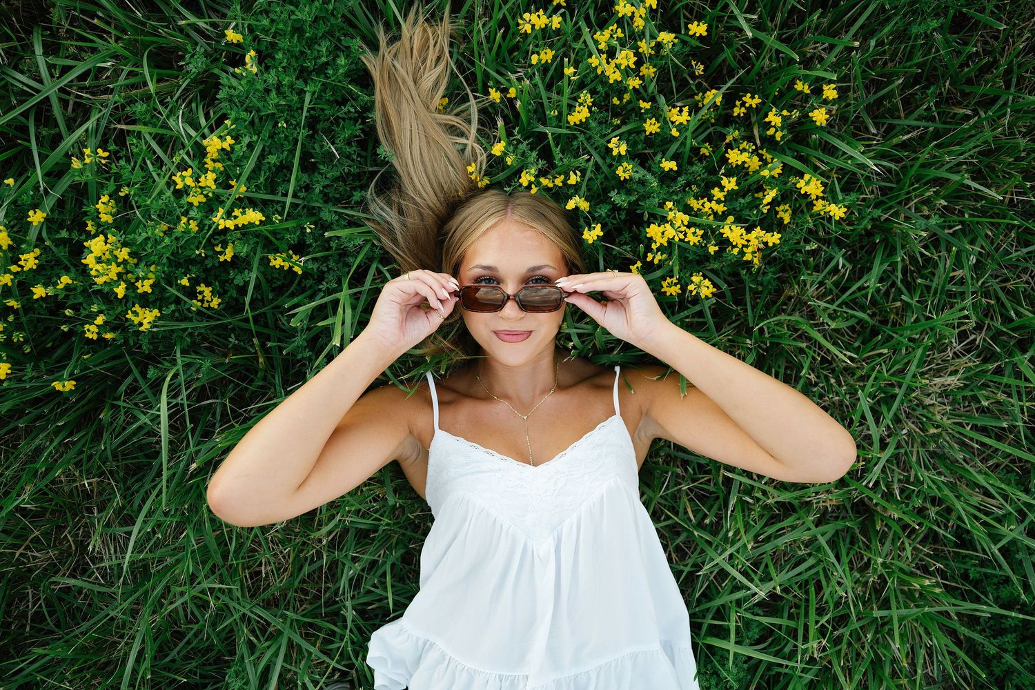 Woman in white dress lying in green grass with yellow flowers, adjusting sunglasses, aerial view posing for Ankeny senior photographer