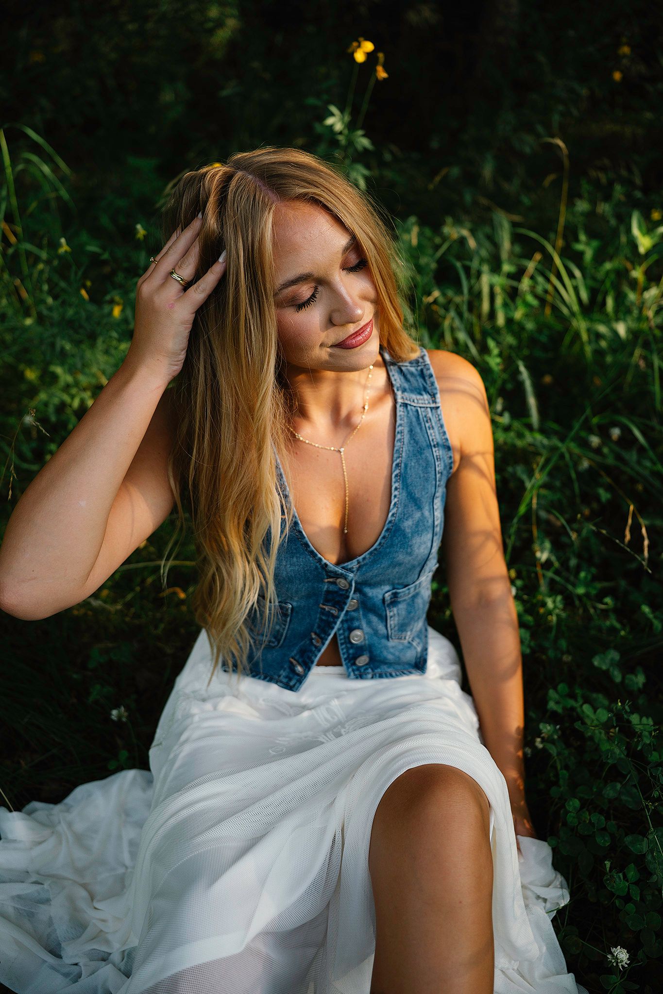 Blonde woman in denim vest and white skirt sits outdoors in lush nature, smiling with eyes closed posing for Des Moines senior photographer