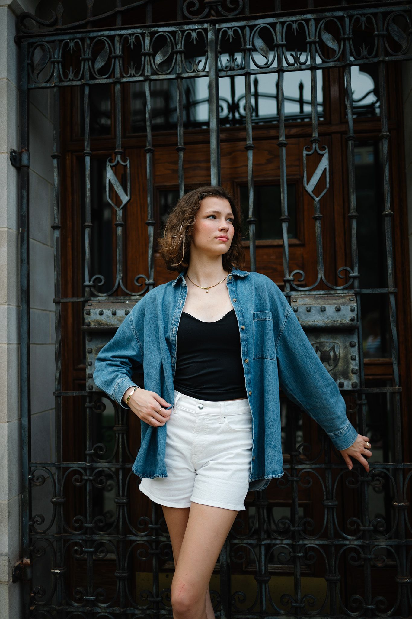 Young woman in denim shirt, black top, and white shorts posing confidently before ornate iron gate.