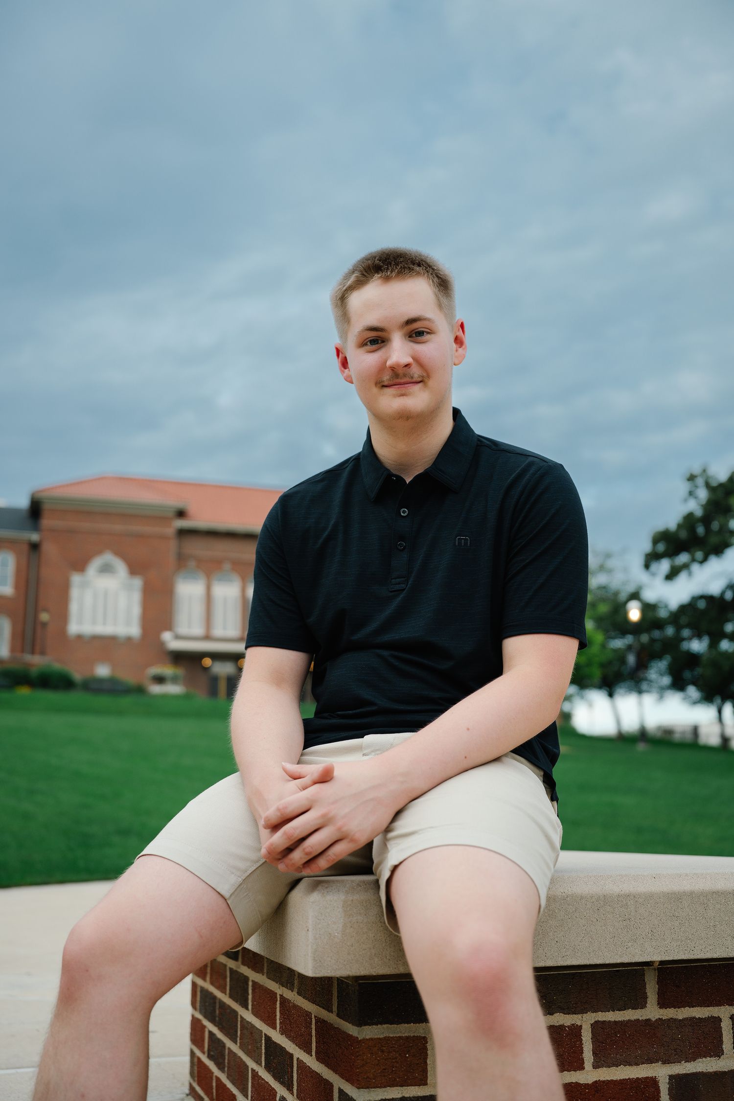 Young man in navy polo and khaki shorts sitting on a brick wall outdoors near a red brick building.