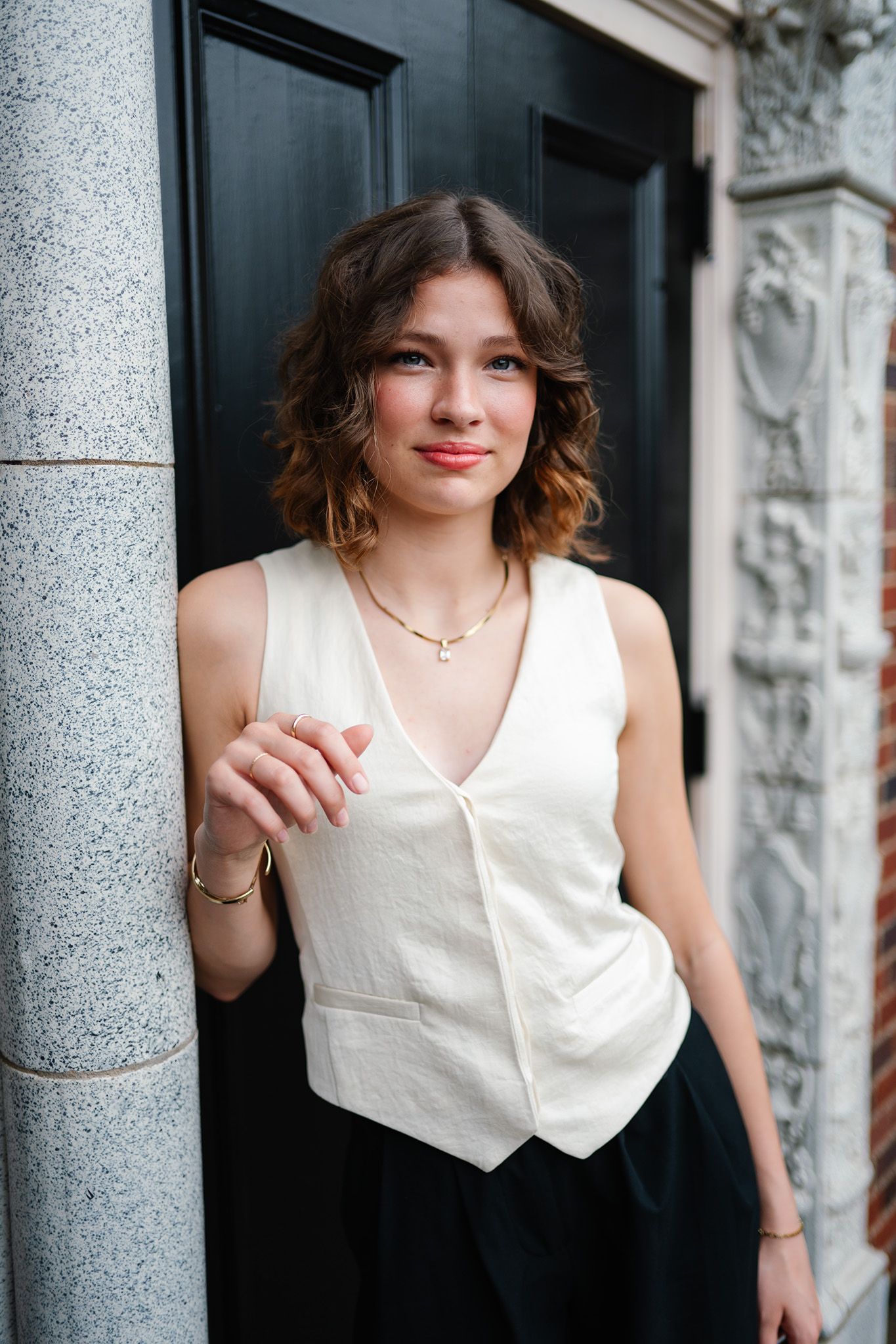 Young woman in white vest top and black skirt leaning against a stone pillar by a black door.