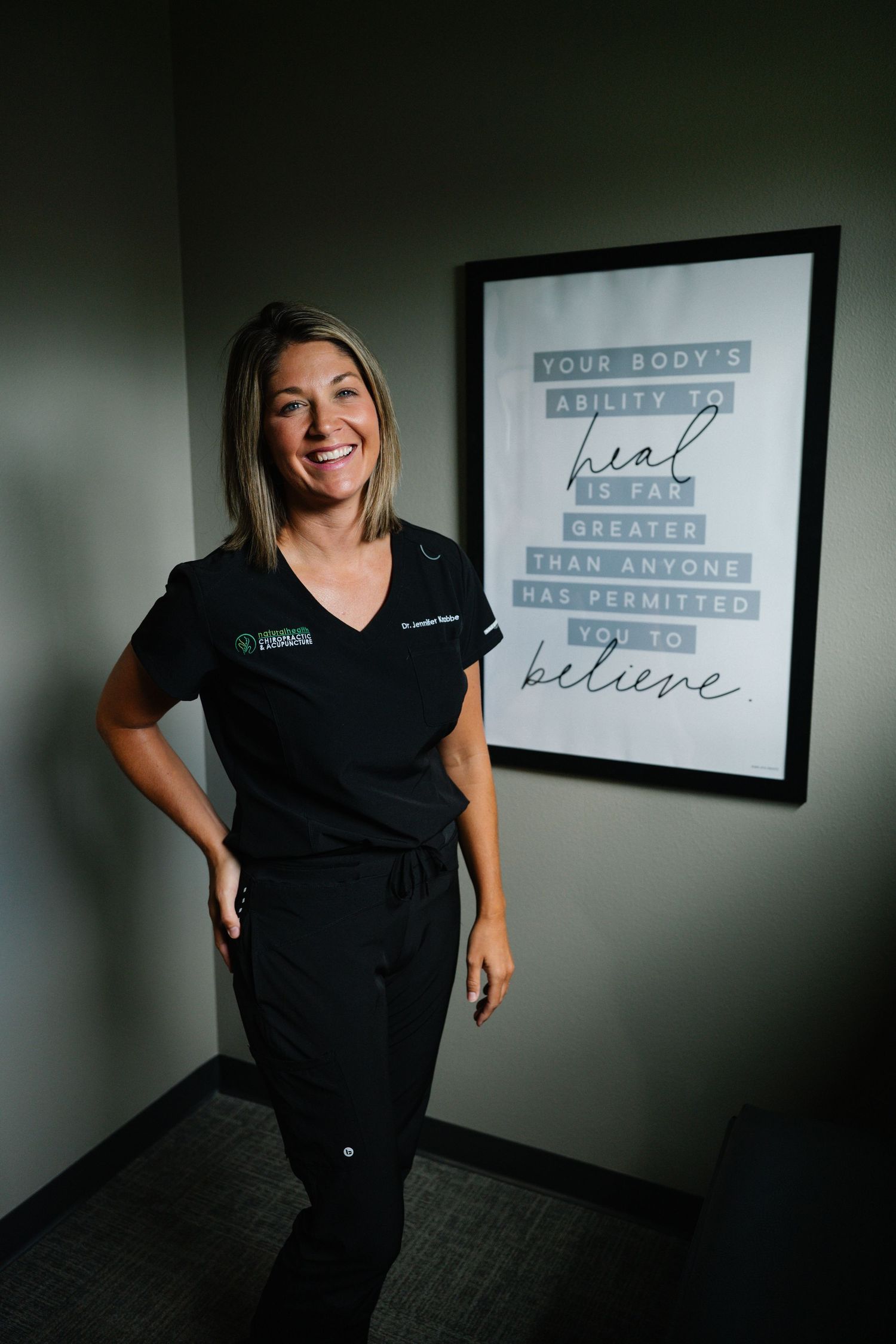 Smiling female healthcare professional in black scrubs standing next to motivational healing quote poster in modern clinic.