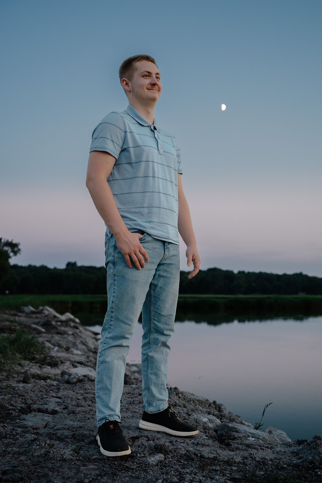 Young man in blue polo shirt and jeans standing on rocky shore by a lake at dusk with moon visible.