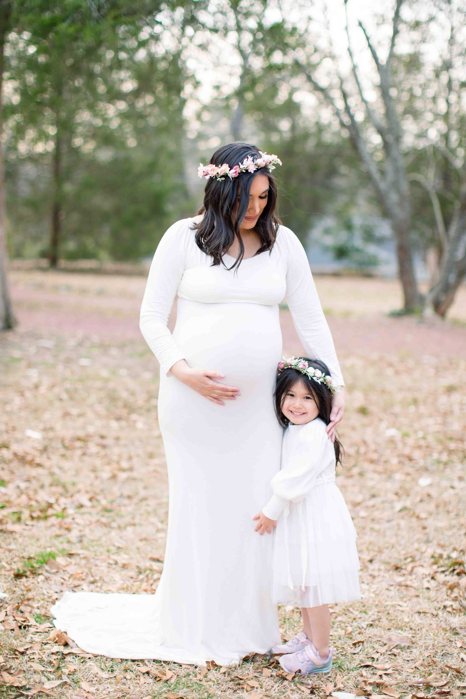 Pregnant mother in white gown with flower crown hugging young daughter outdoors.