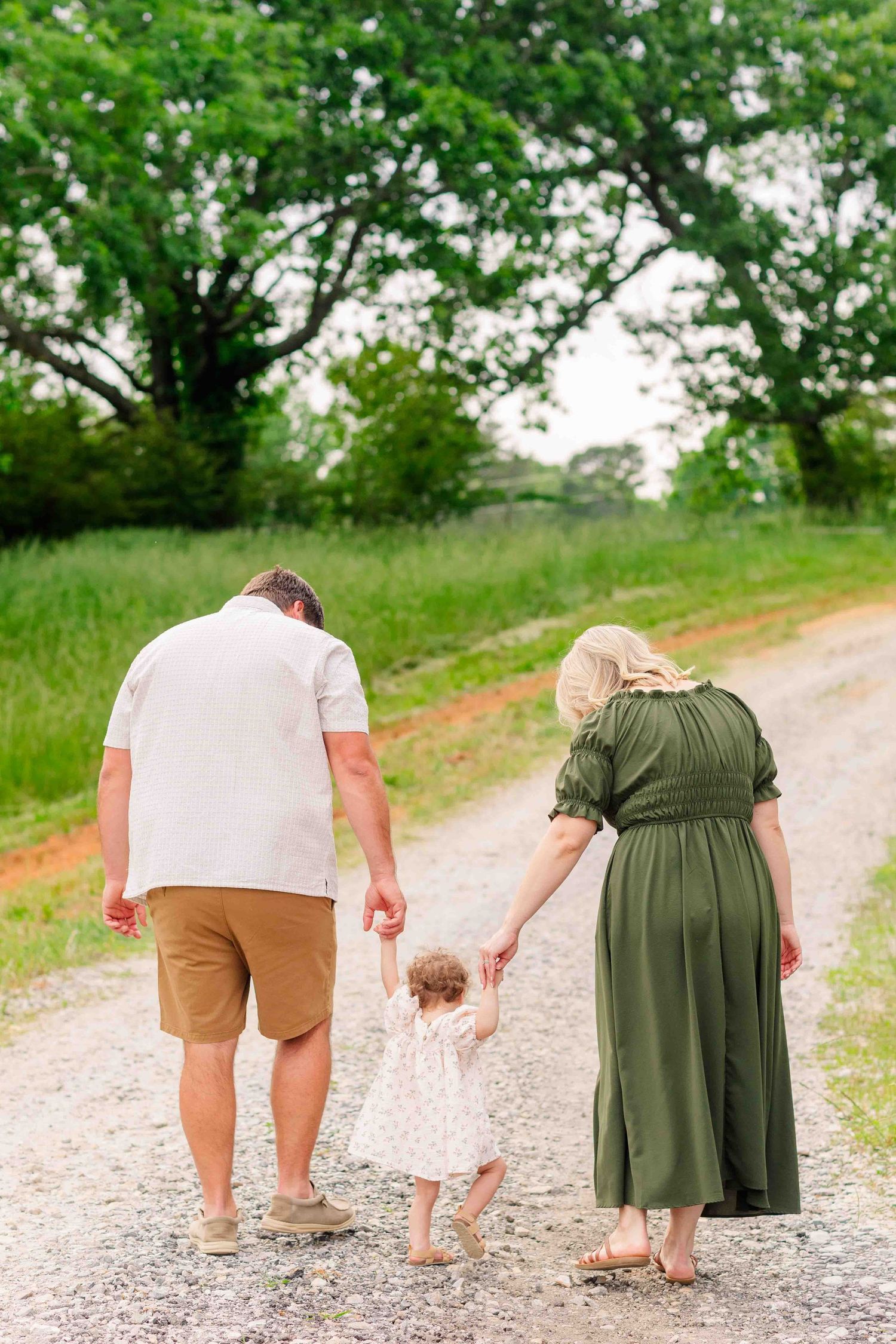 Family of three walking hand-in-hand down a rustic gravel path surrounded by green fields.