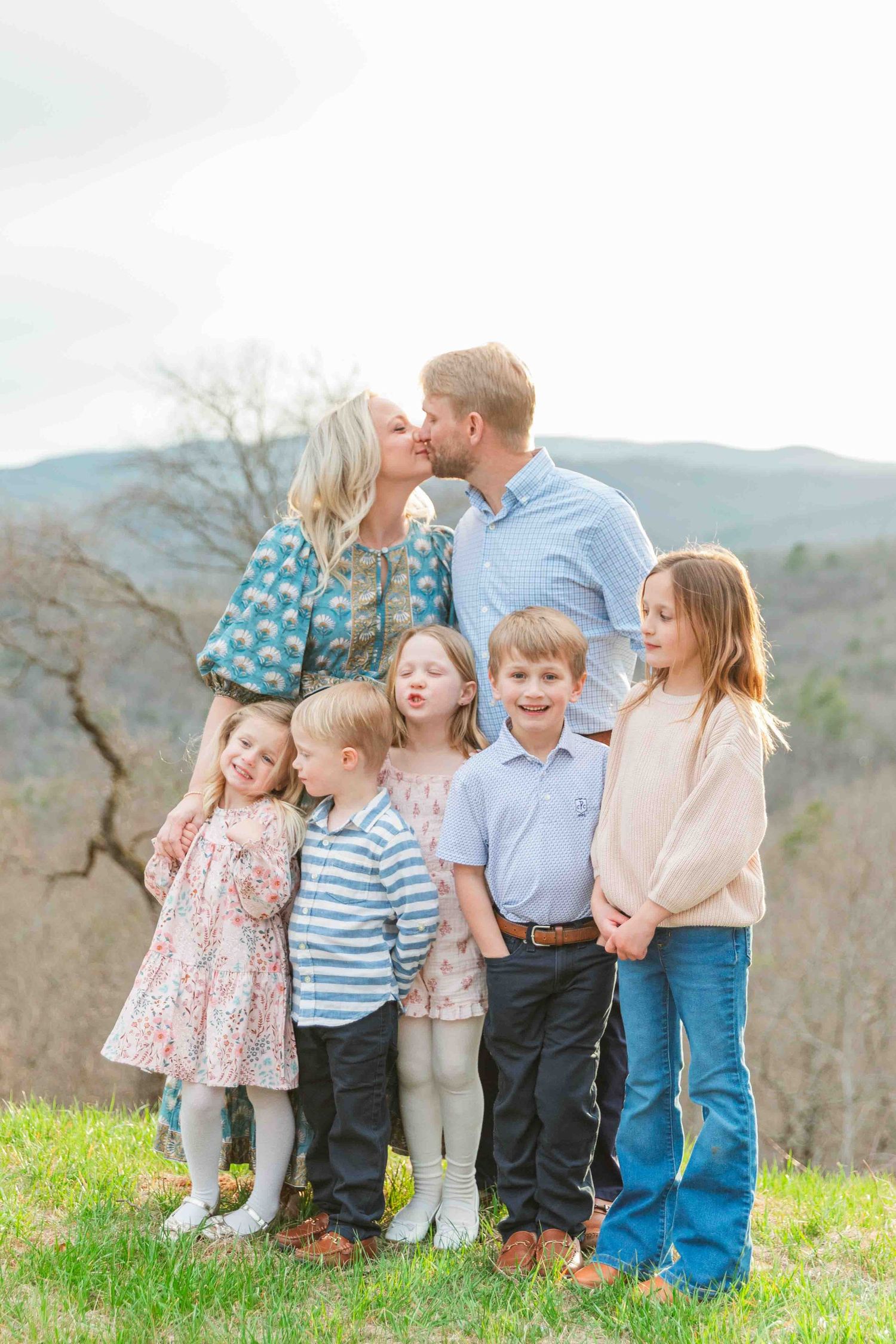 Family of seven with five children posing on a grassy hill with mountain views while parents kiss.