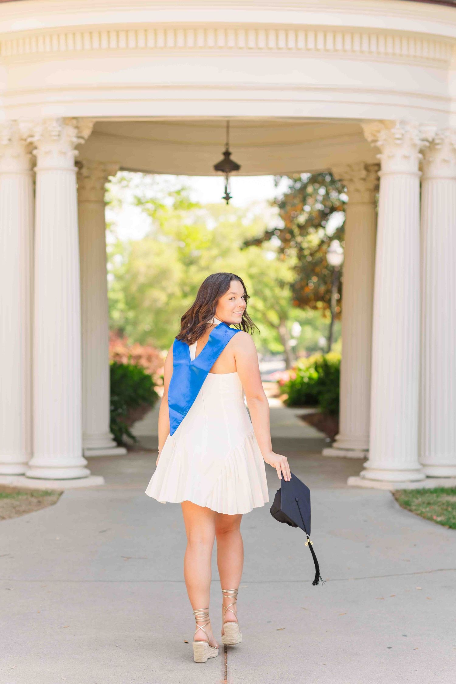 Brunette graduate in white dress with blue stole walks through white columned pavilion holding cap.