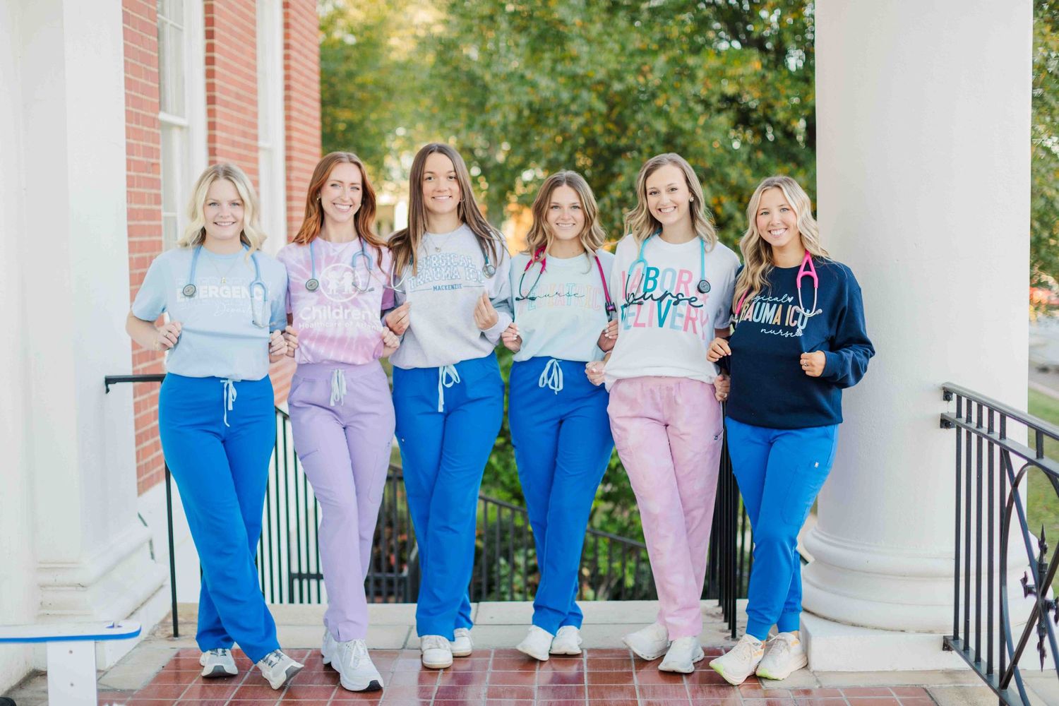 Six nursing students in scrubs smile while holding certificates outside a brick building.