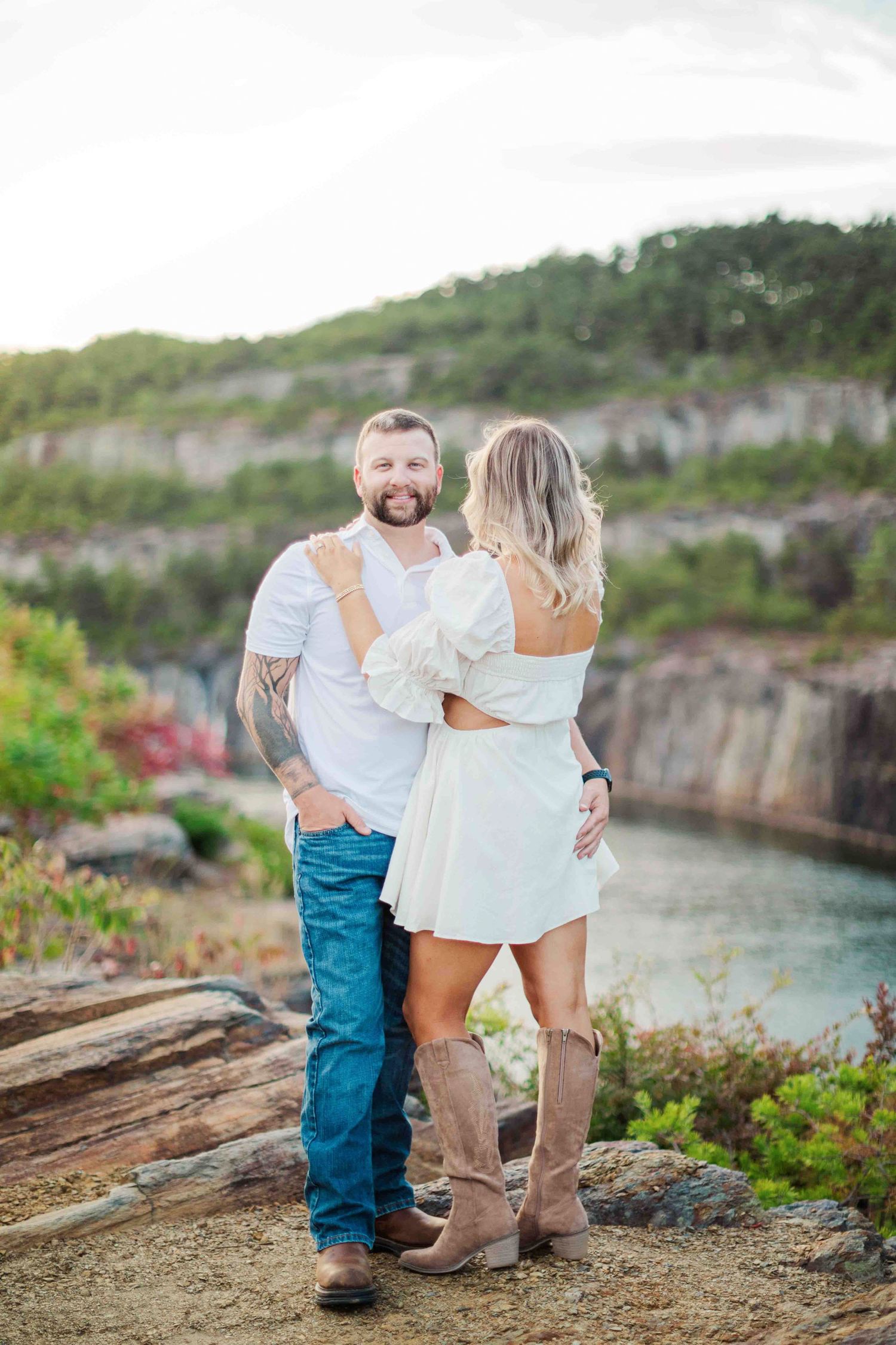 Couple embracing on rocky cliff overlooking scenic quarry lake at sunset, woman in white dress and cowboy boots.