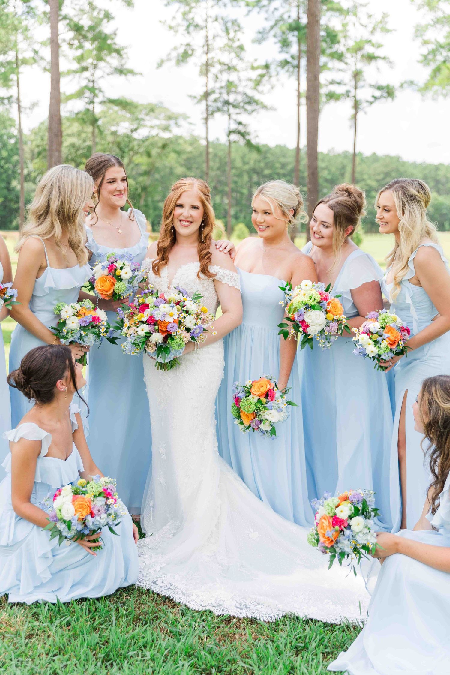 Bride in lace gown surrounded by bridesmaids in light blue dresses holding colorful bouquets.
