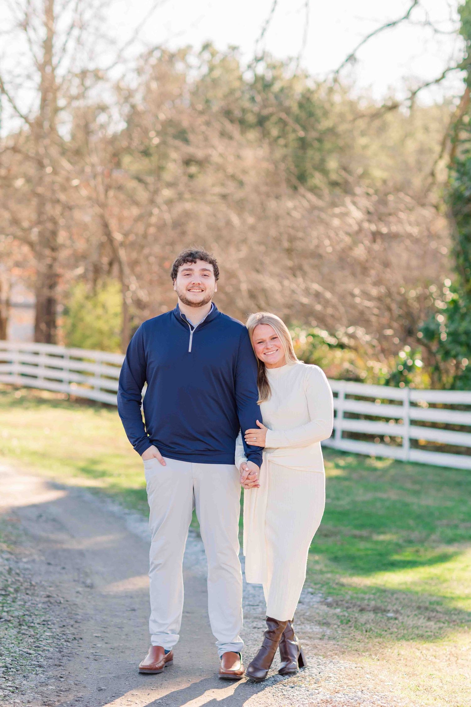 Smiling couple standing together on a gravel path with white fence and autumn trees in background.