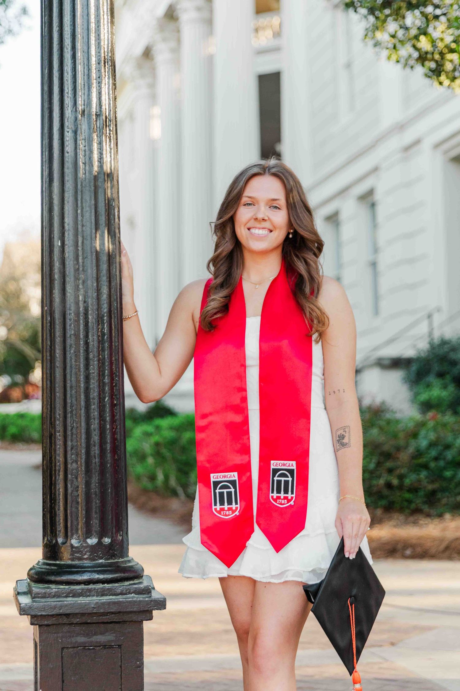 Smiling graduate in white dress with red university stole holds cap beside lamppost on campus.
