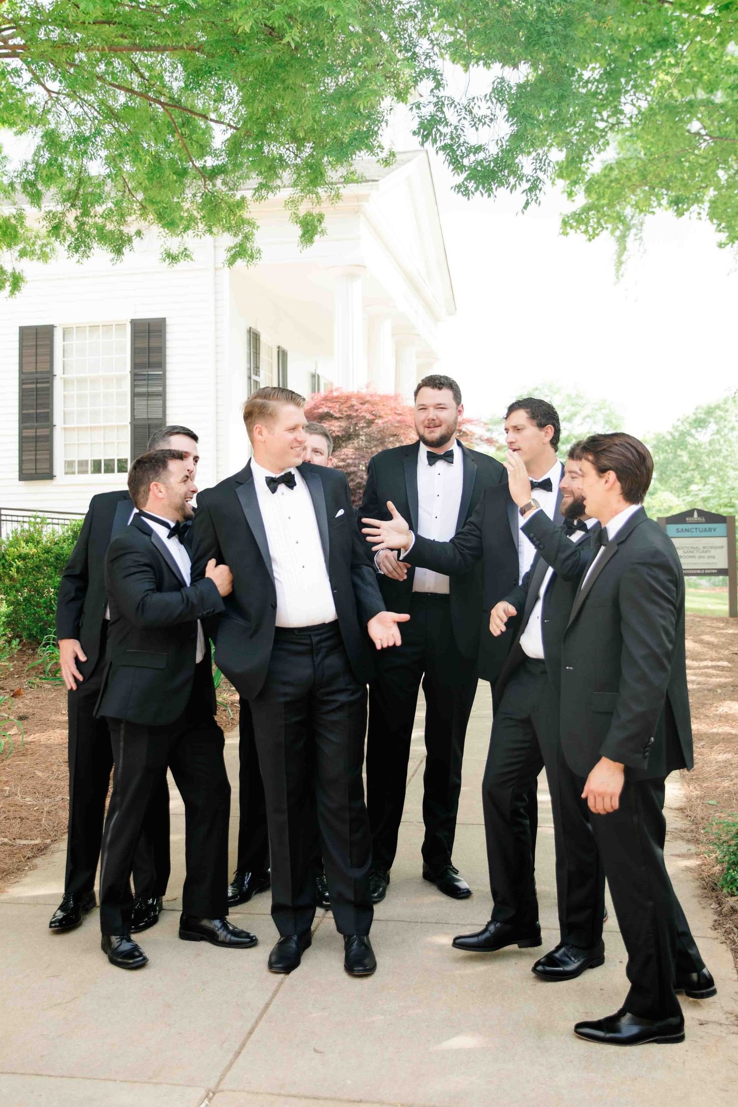 Groomsmen in black tuxedos gathered in lively conversation outside elegant white venue.
