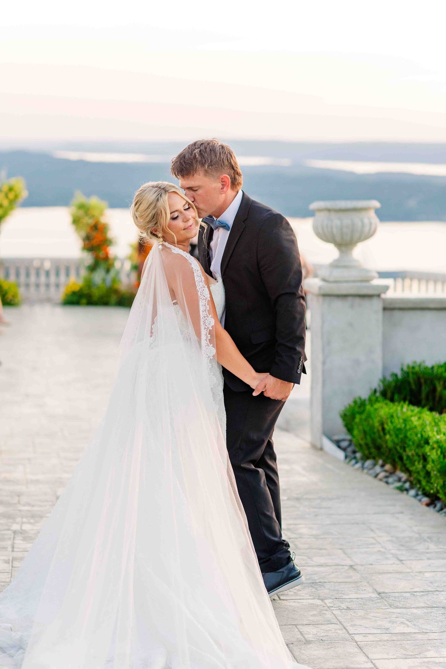 Bride and groom share a romantic kiss on a terrace overlooking a lake at golden hour during their wedding.