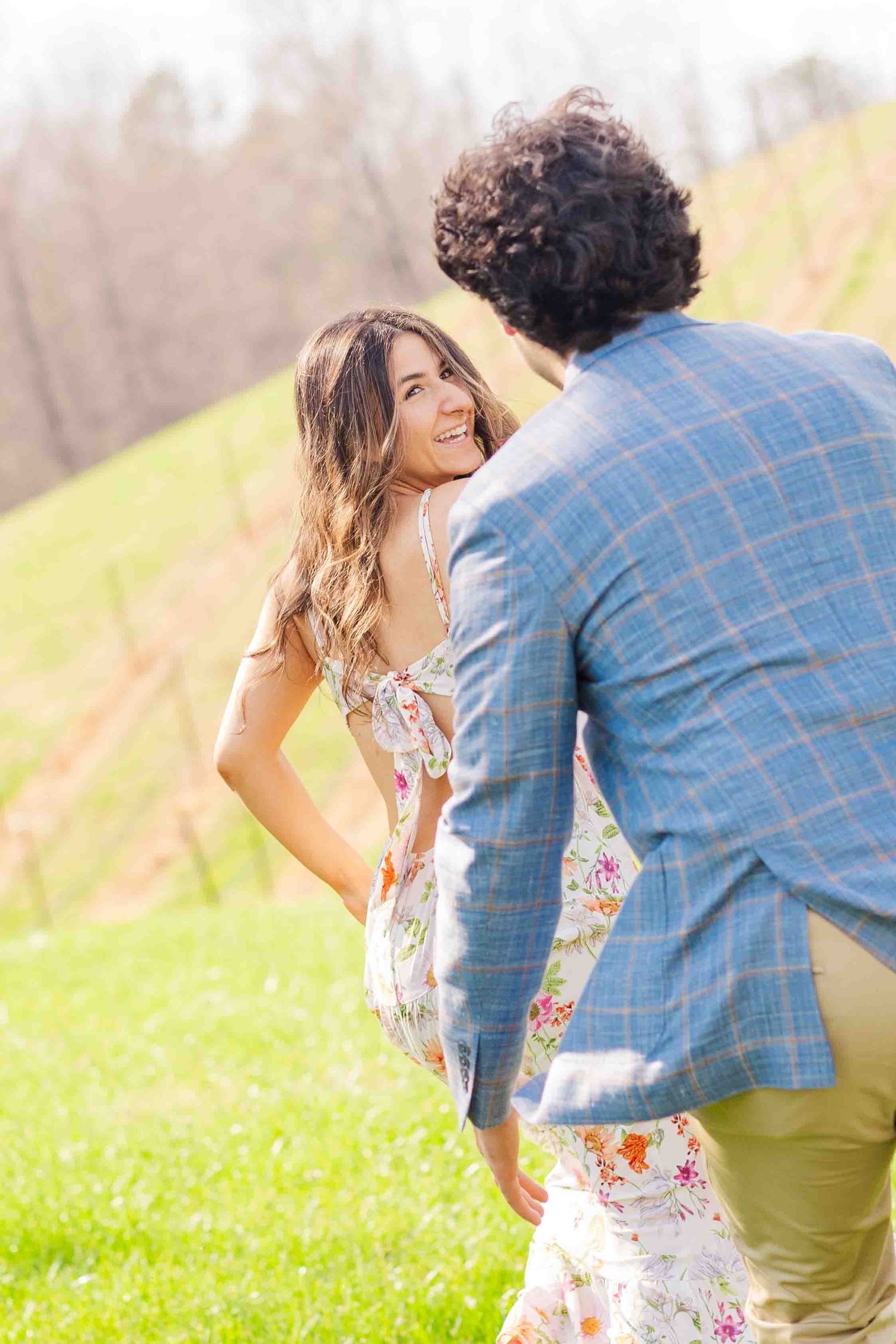 Woman in floral dress smiling over shoulder at curly-haired man in plaid blazer in vineyard setting.