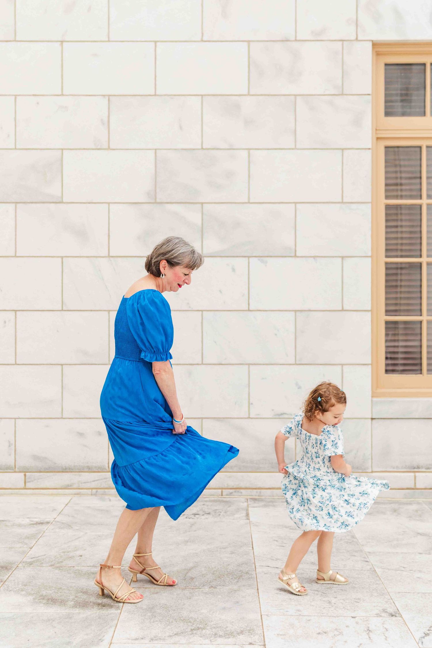 Grandmother in blue dress twirling with young granddaughter in floral dress against marble wall.
