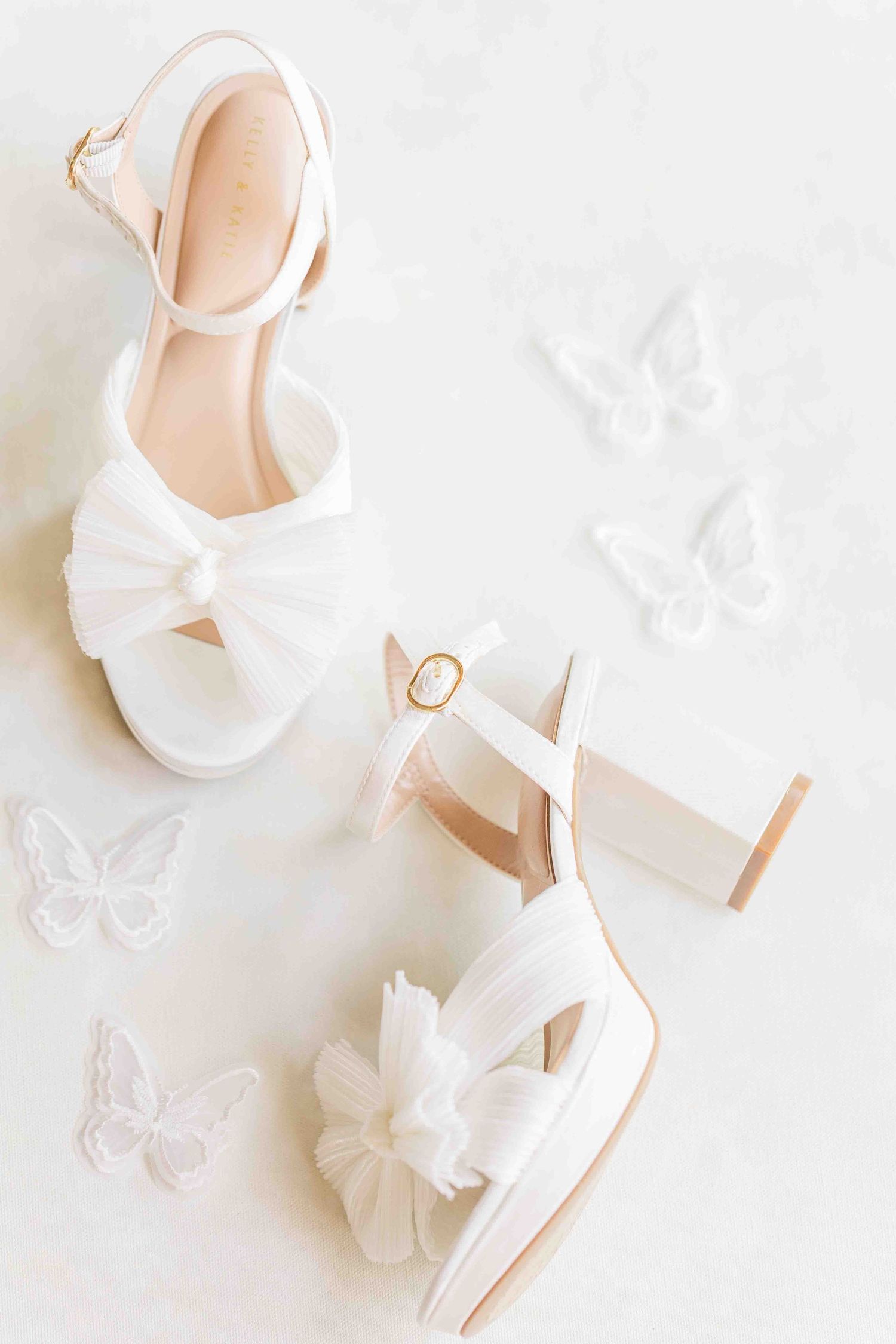 White bridal heels with bow detail displayed on a light surface surrounded by delicate white butterfly decorations.