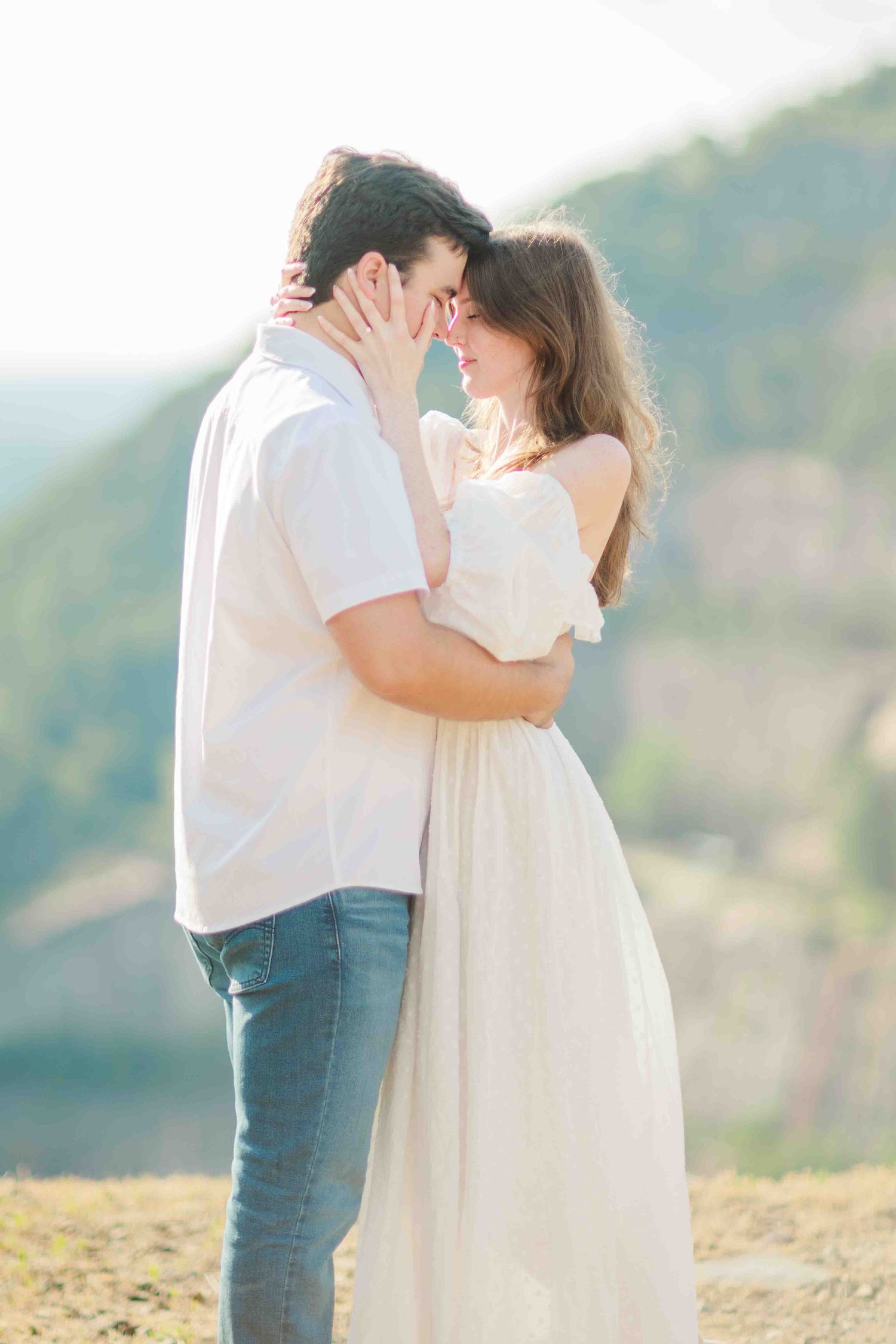 Romantic couple nearly kissing outdoors with mountain landscape in background, both wearing white.