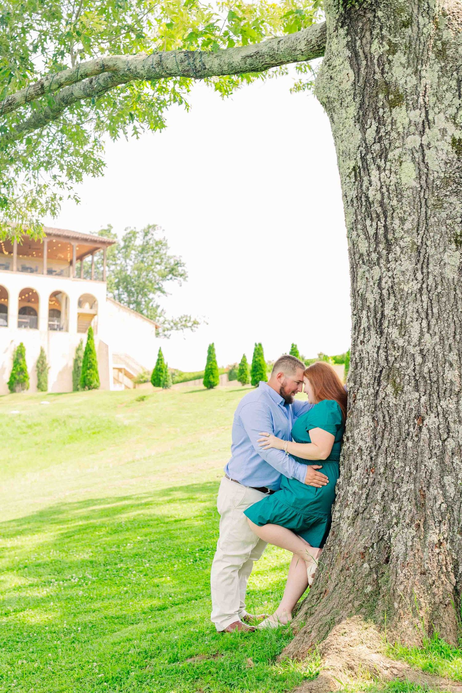 Couple sharing romantic moment leaning against large tree trunk on green lawn near elegant winery building.