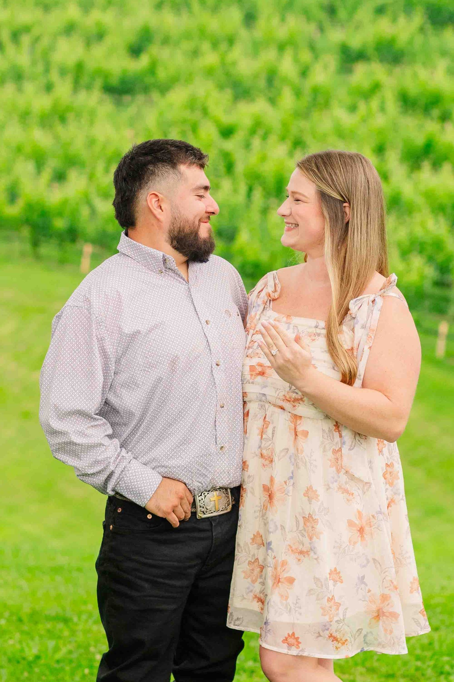 Happy couple smiling at each other in front of lush green vineyard, woman in floral dress and man in striped shirt.