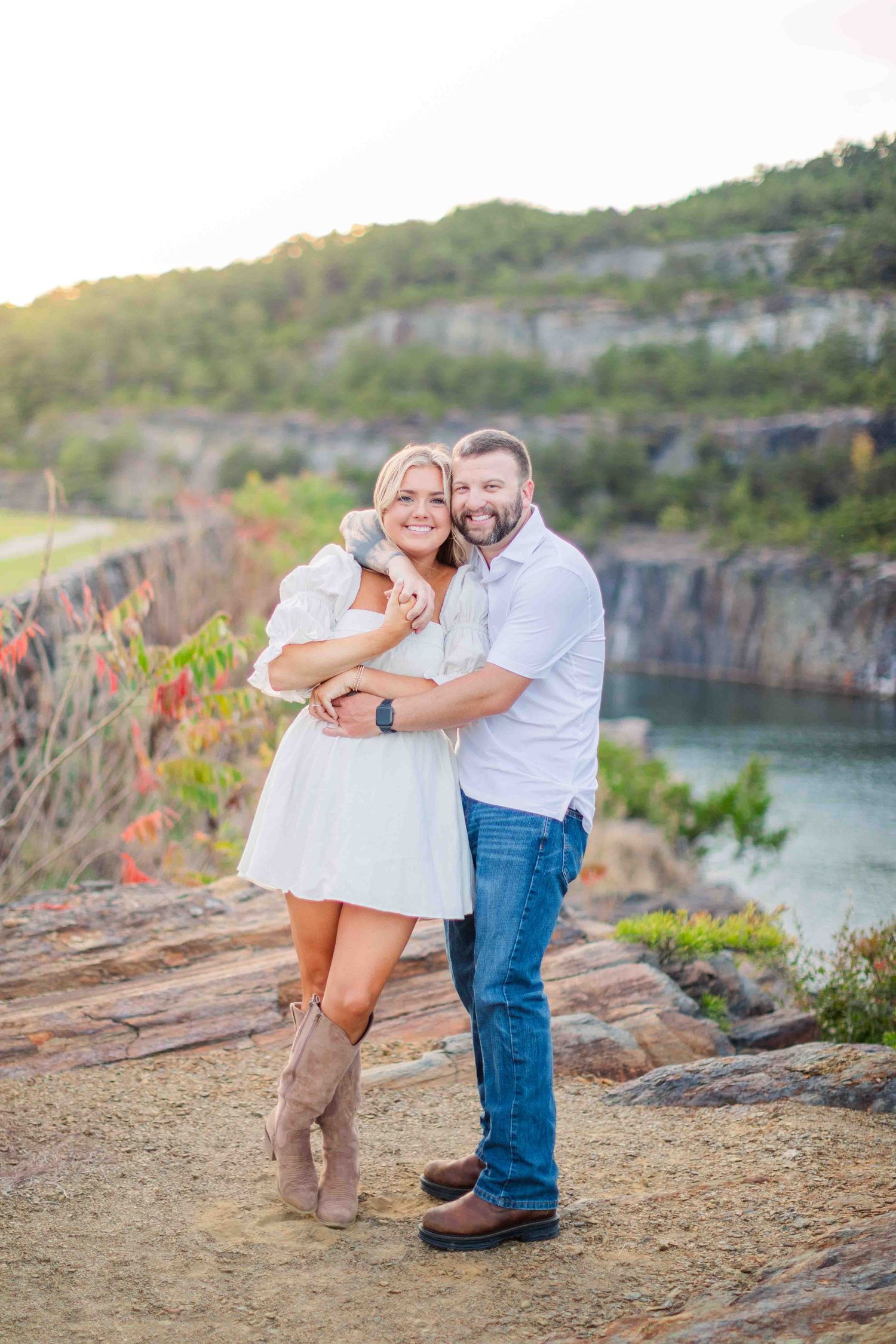 Happy couple hugging and smiling at scenic rocky quarry overlook at golden hour, woman in white dress and boots.