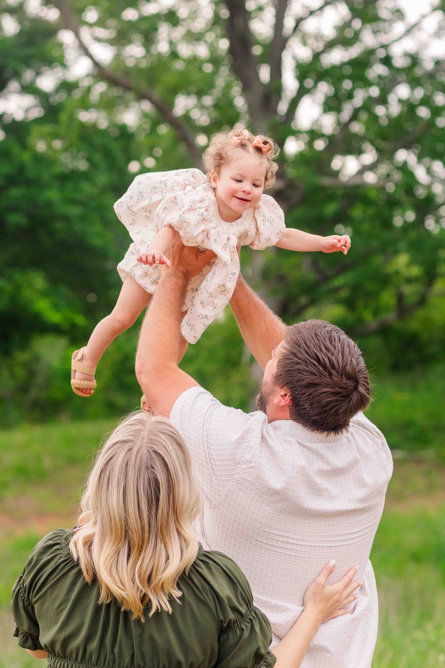 Parents joyfully lifting their laughing toddler daughter into the air in a lush green park.