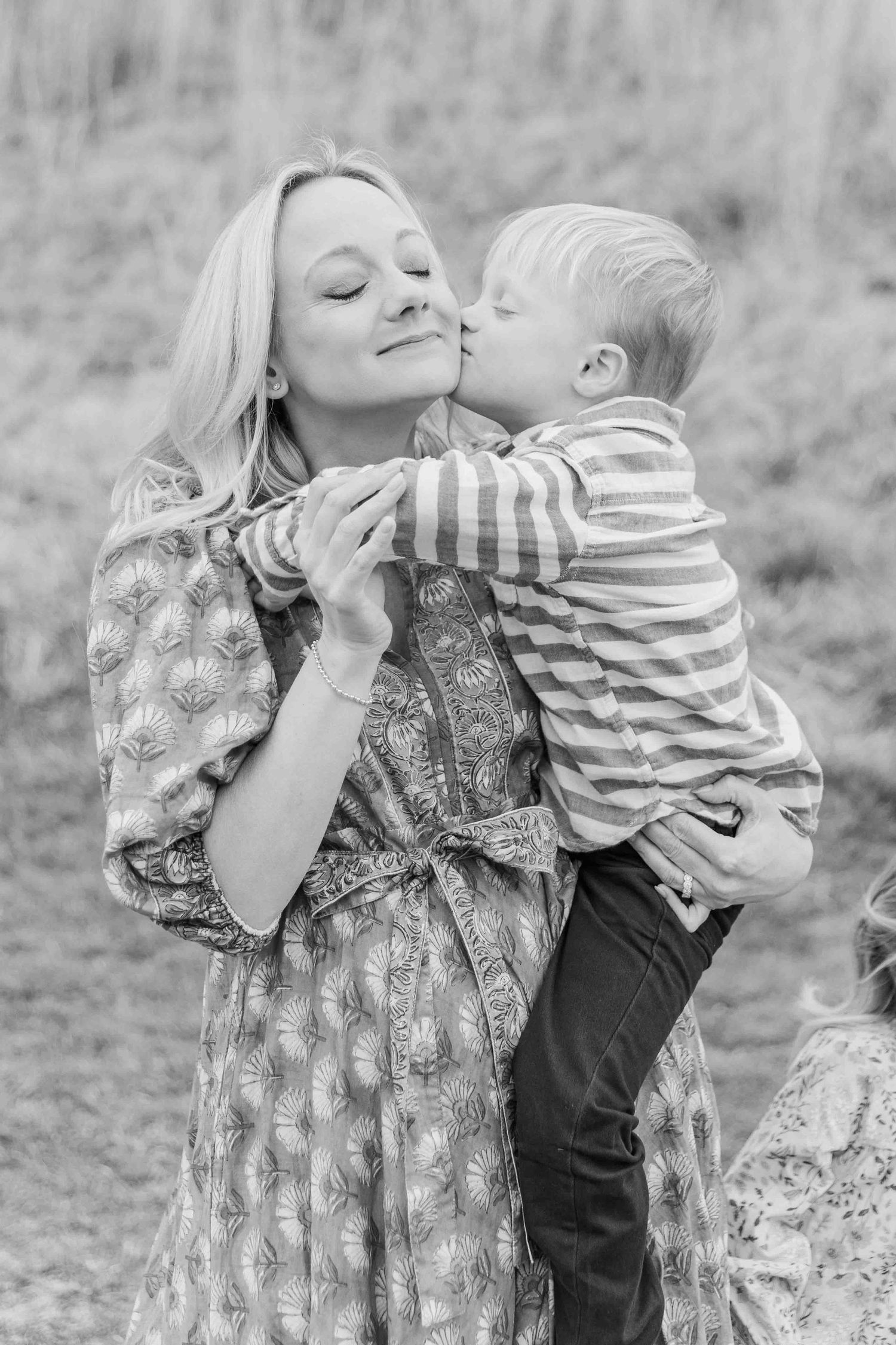 Black and white portrait of a smiling mother holding her young son as he kisses her cheek outdoors.