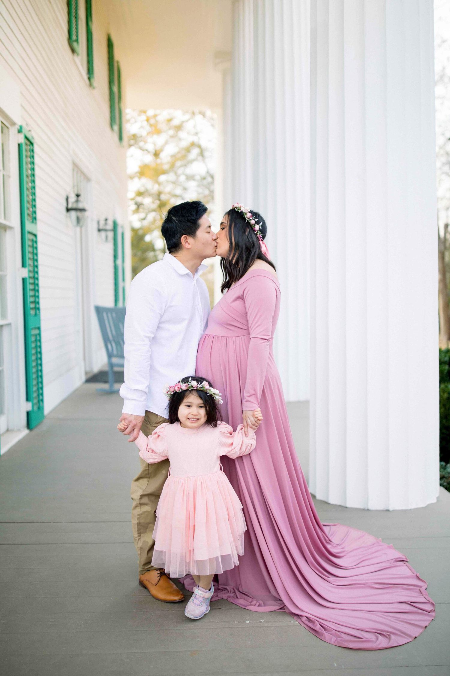 Expecting couple kissing on porch in pink and white outfits with young daughter.
