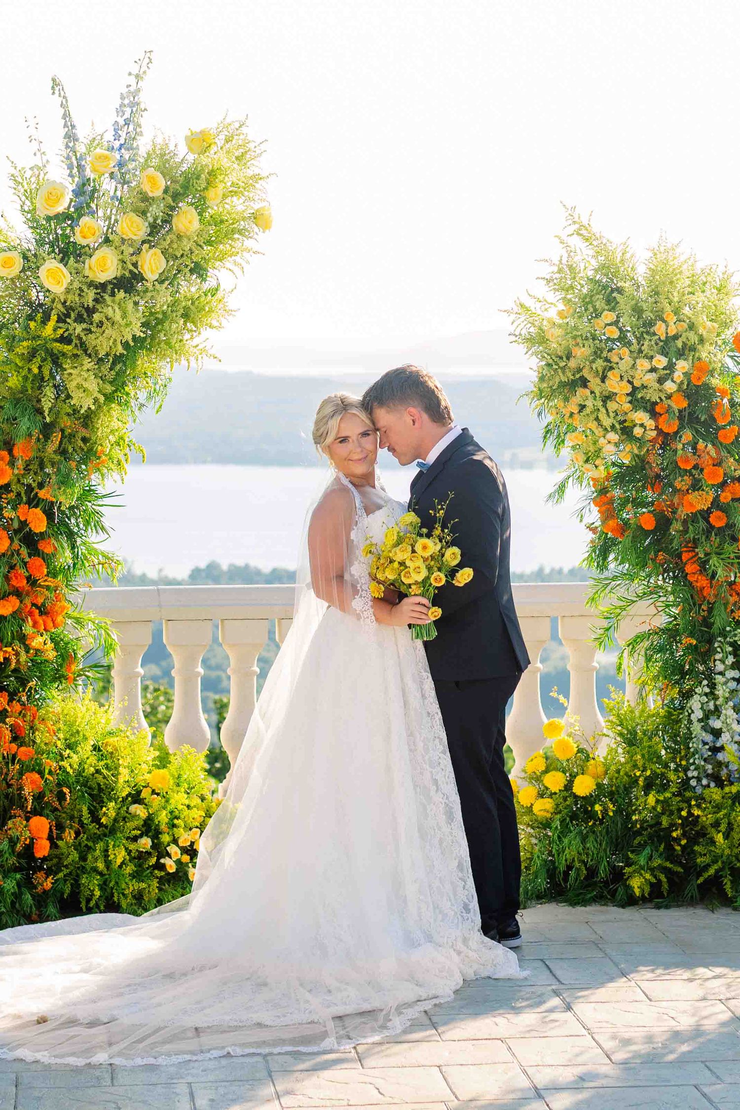Bride and groom embrace under floral arch with yellow and orange flowers, overlooking scenic waterfront at outdoor wedding ceremony.