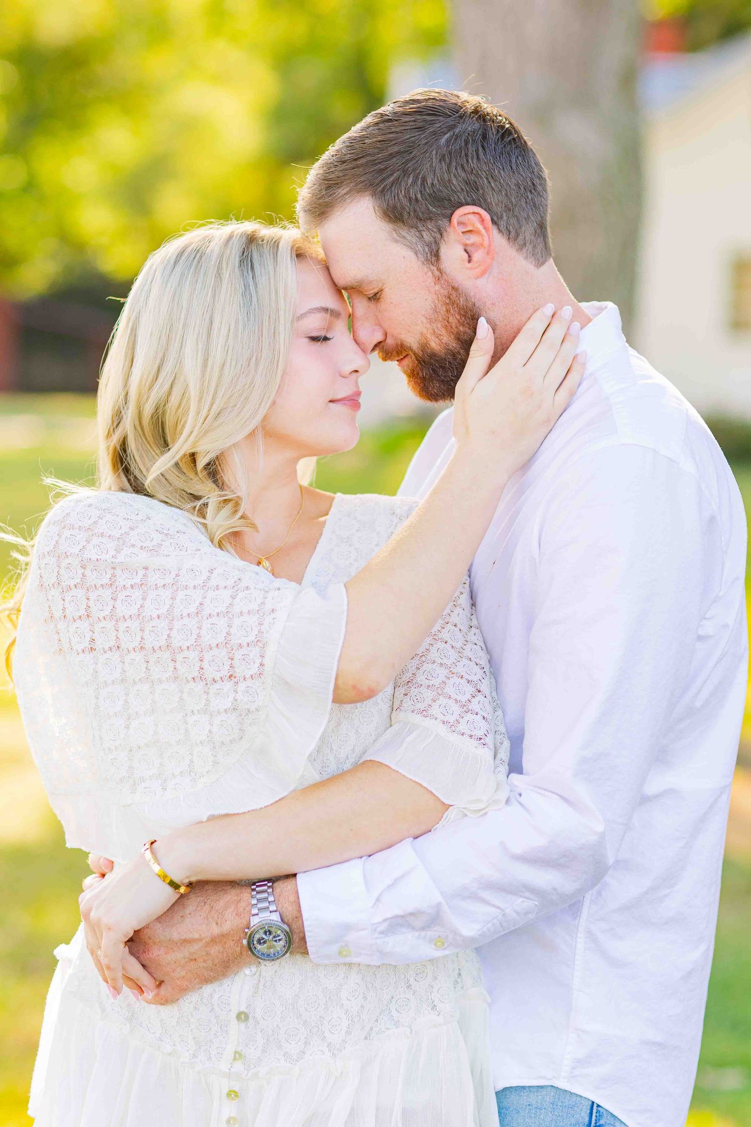 Couple embracing forehead-to-forehead in golden sunlight, woman in white lace dress holding mans face.