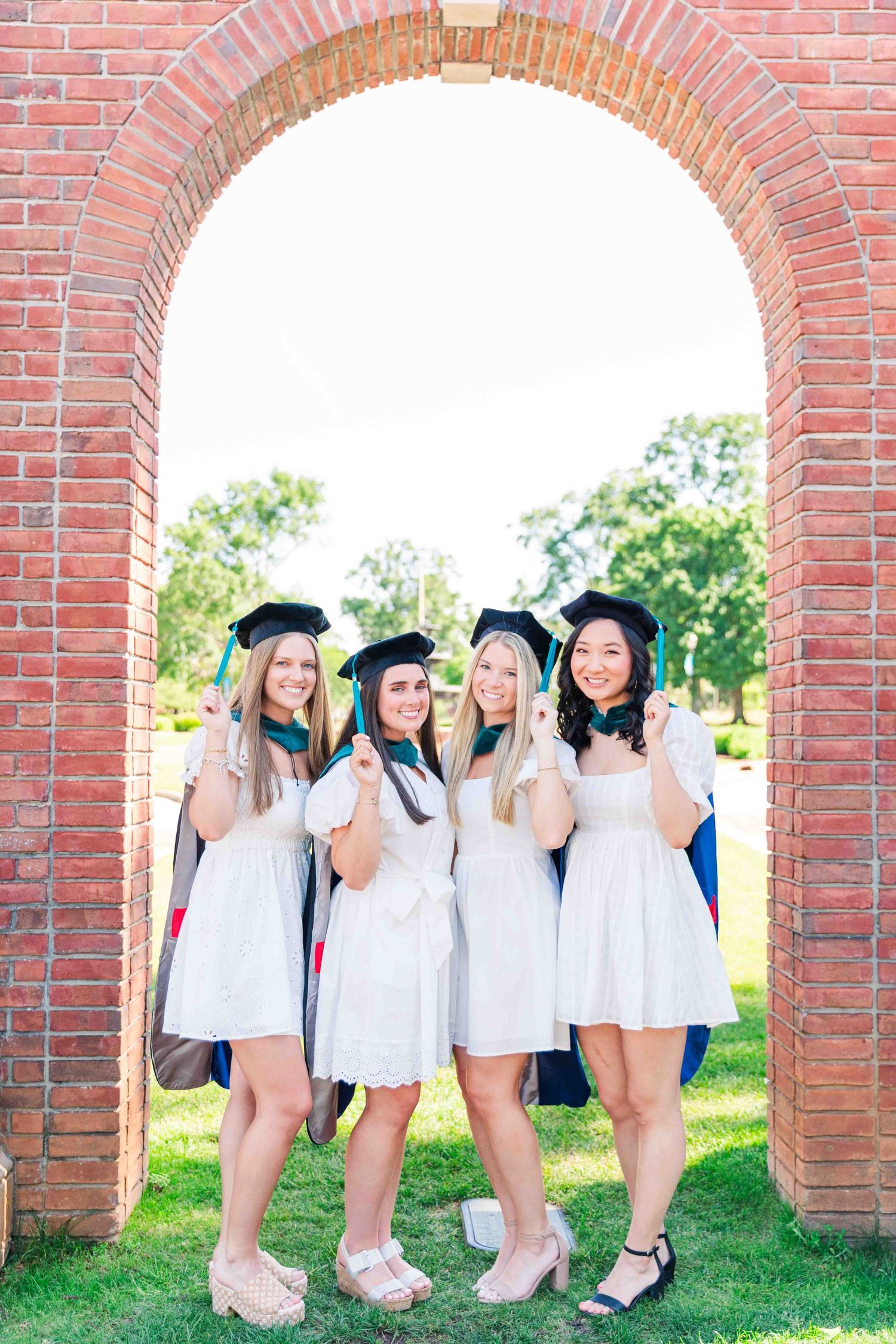 Four graduates in white dresses and black caps pose under brick campus archway.