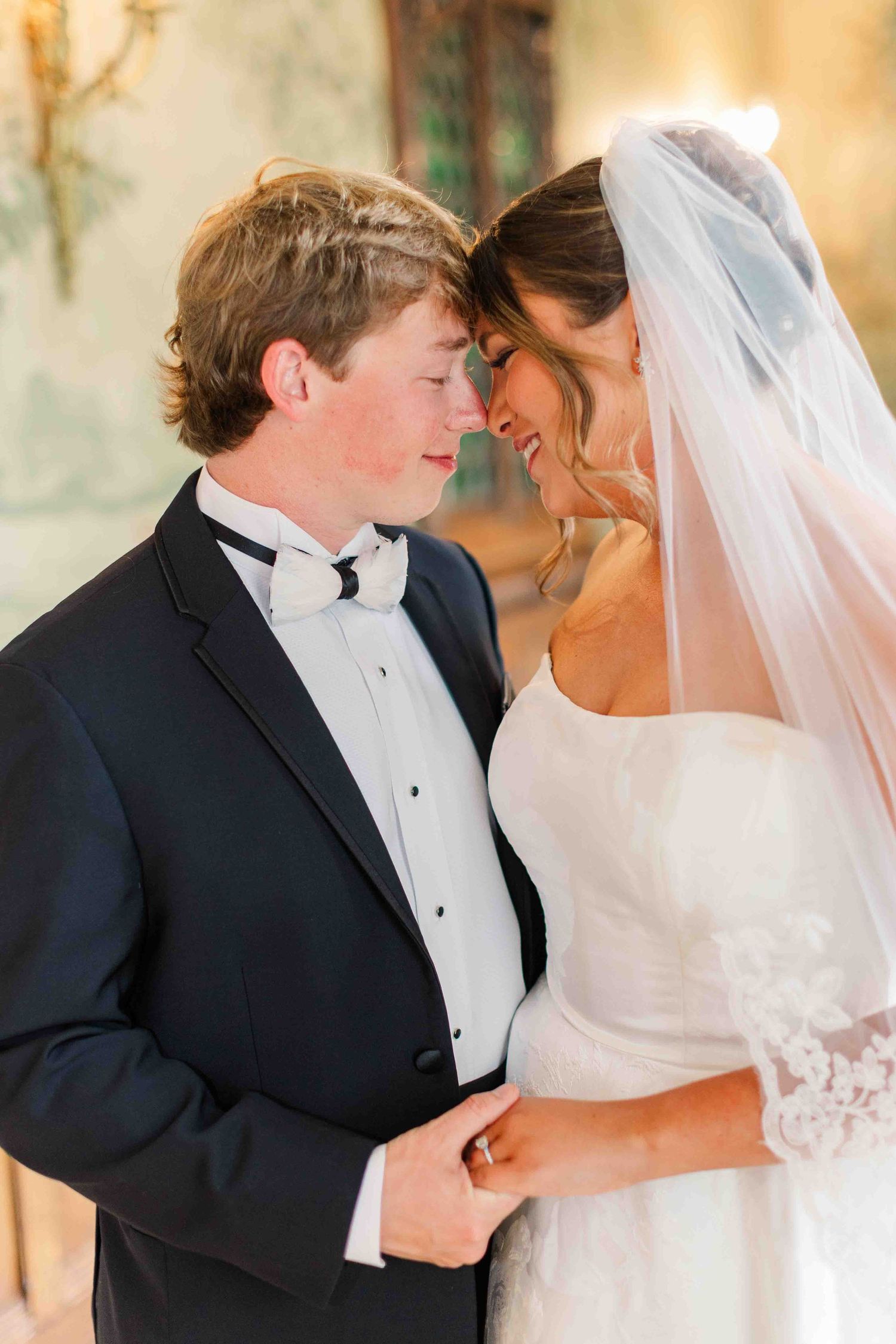 A bride and groom share an intimate moment, touching foreheads, dressed in wedding attire with veil and tuxedo.