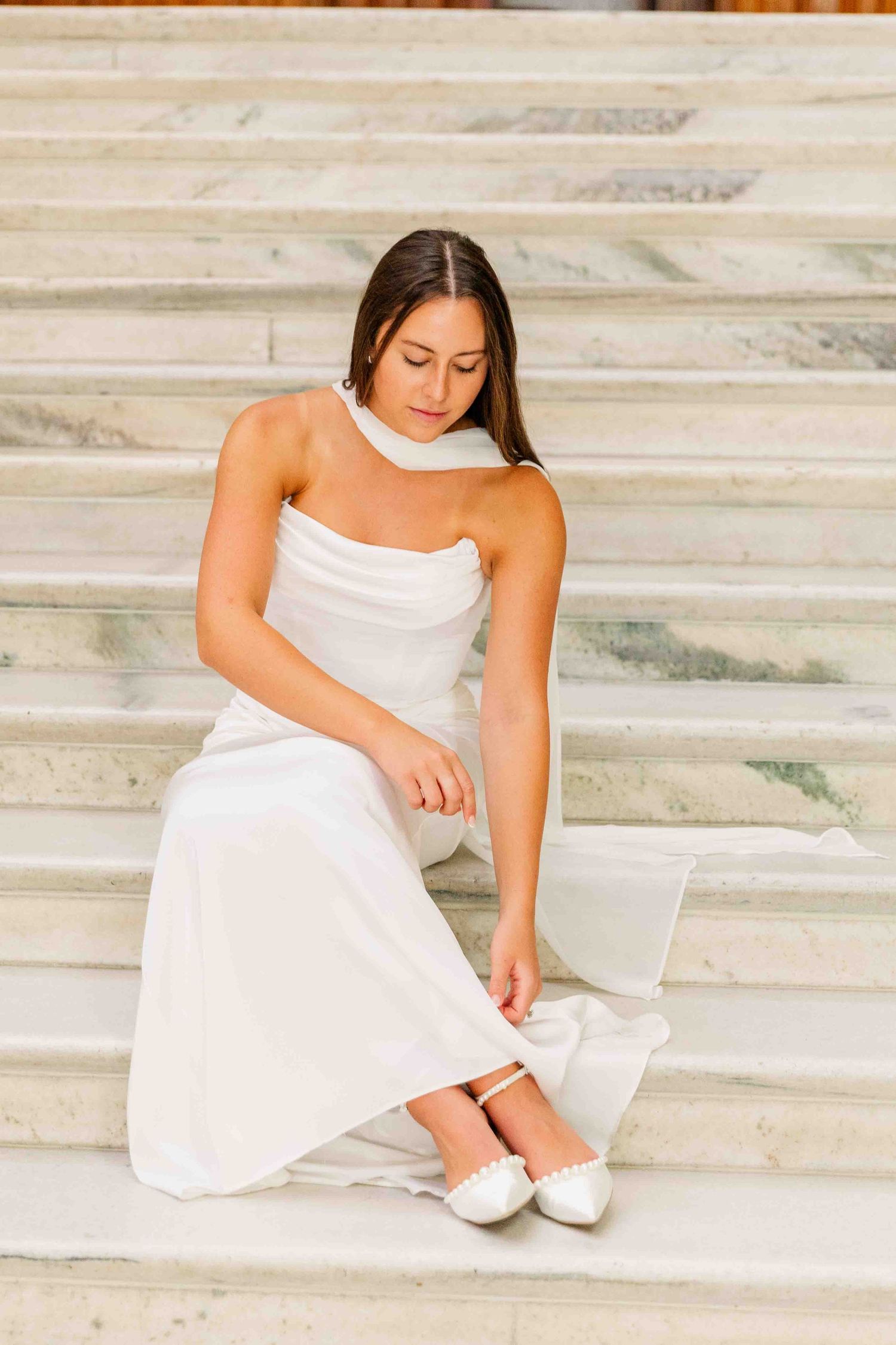 Bride in white gown sitting on marble steps, adjusting her white flat shoes, with elegant draping detail.